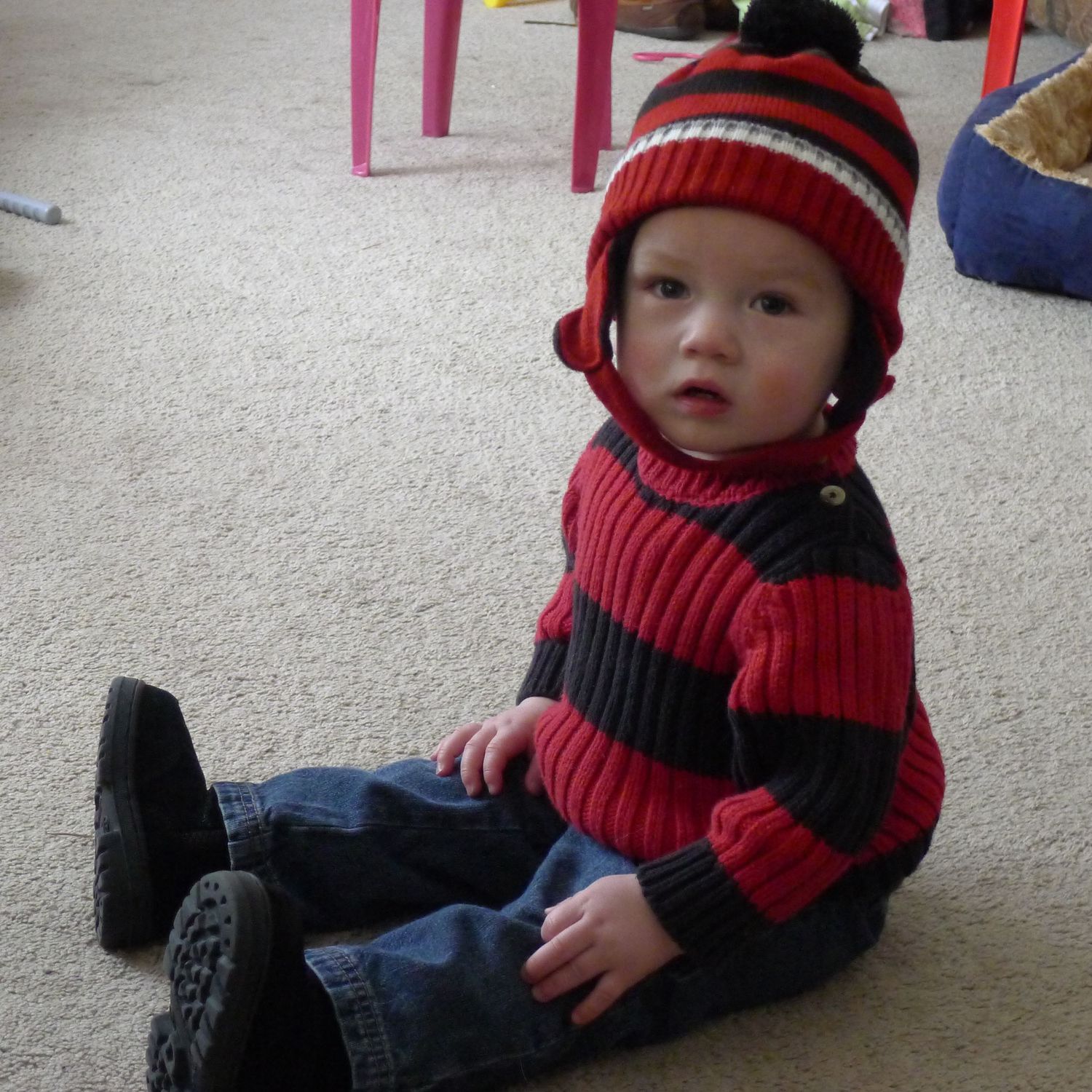 A baby wearing a red striped sweater and matching hat sits on carpet in jeans and black shoes.
