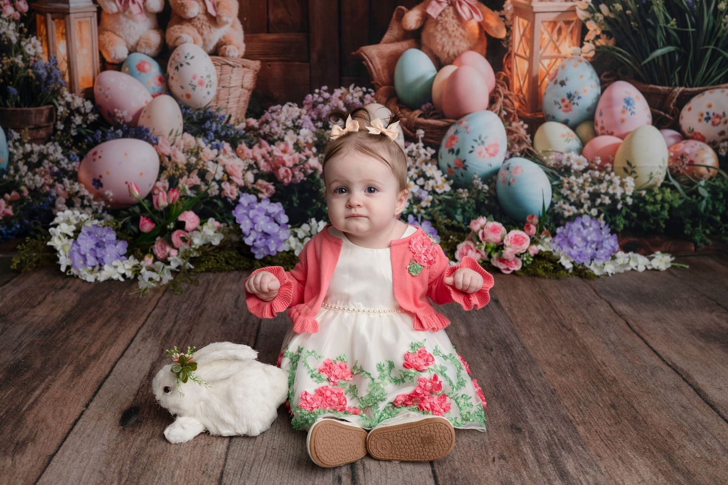 Baby girl in pink floral dress sits on wood floor with white bunny and colorful Easter eggs and flowers.