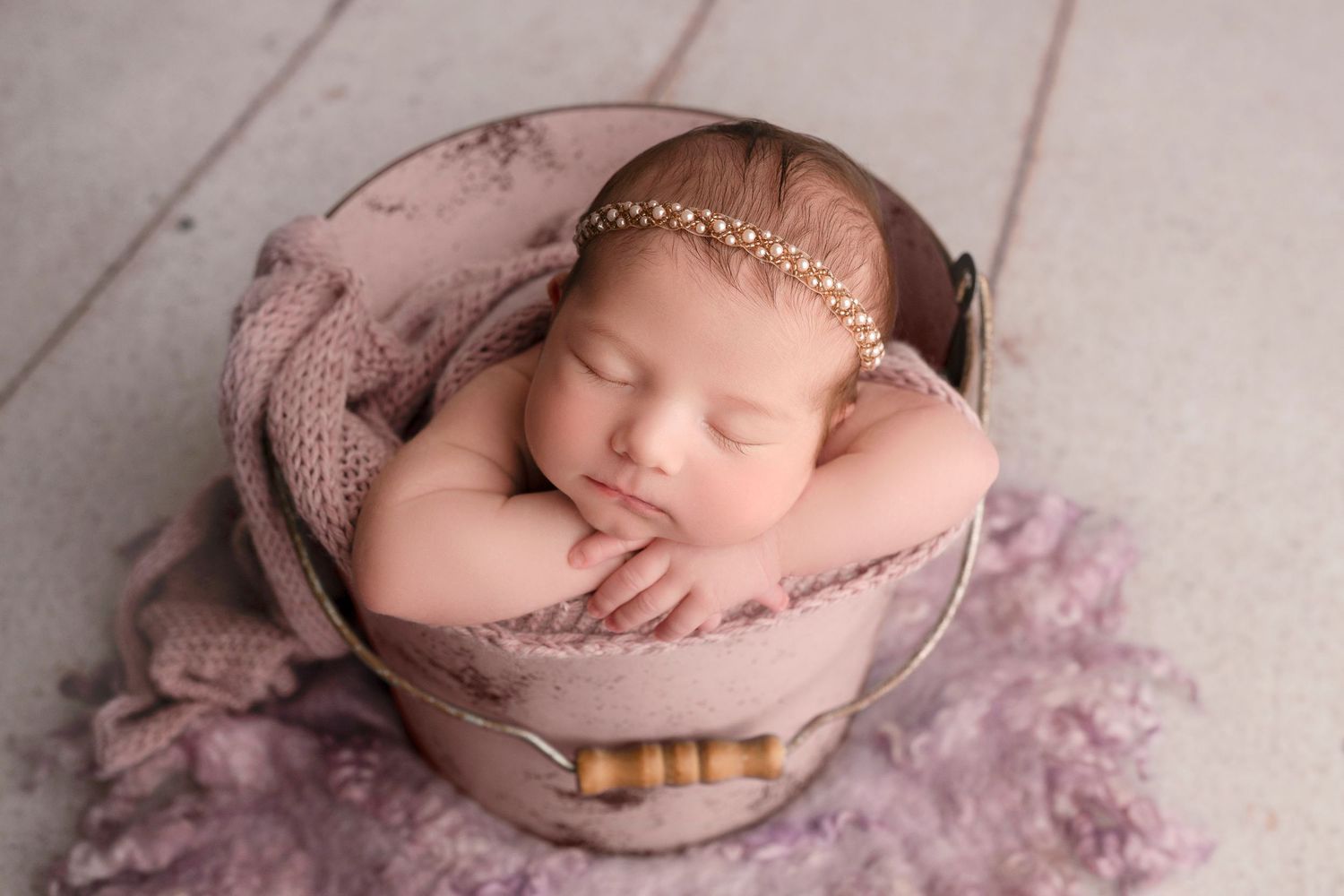 Newborn baby girl sleeping peacefully in a vintage pink bucket with a gold pearl headband and soft purple blanket.