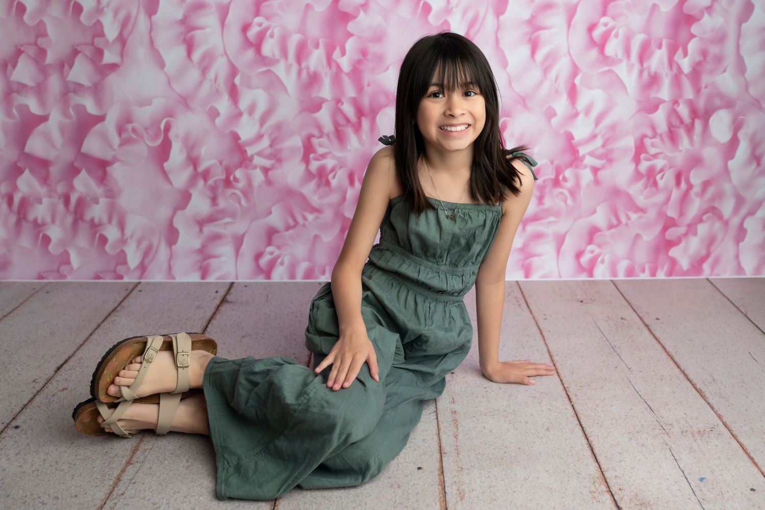 Young girl in green dress sits smiling on white wood floor against pink floral backdrop.