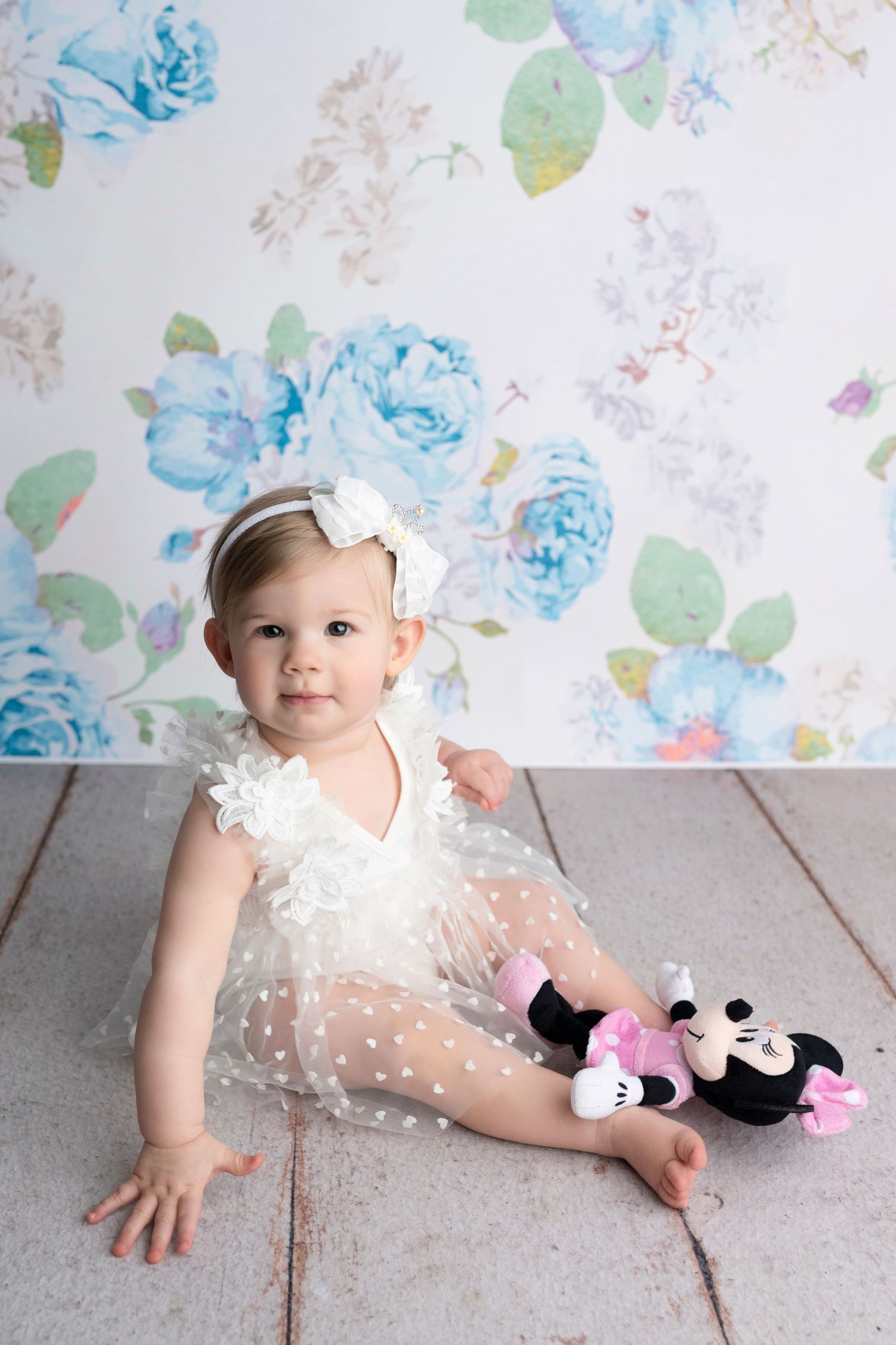 Baby girl in white tutu dress with bow headband sits on wood floor holding Mickey Mouse toy before floral backdrop.