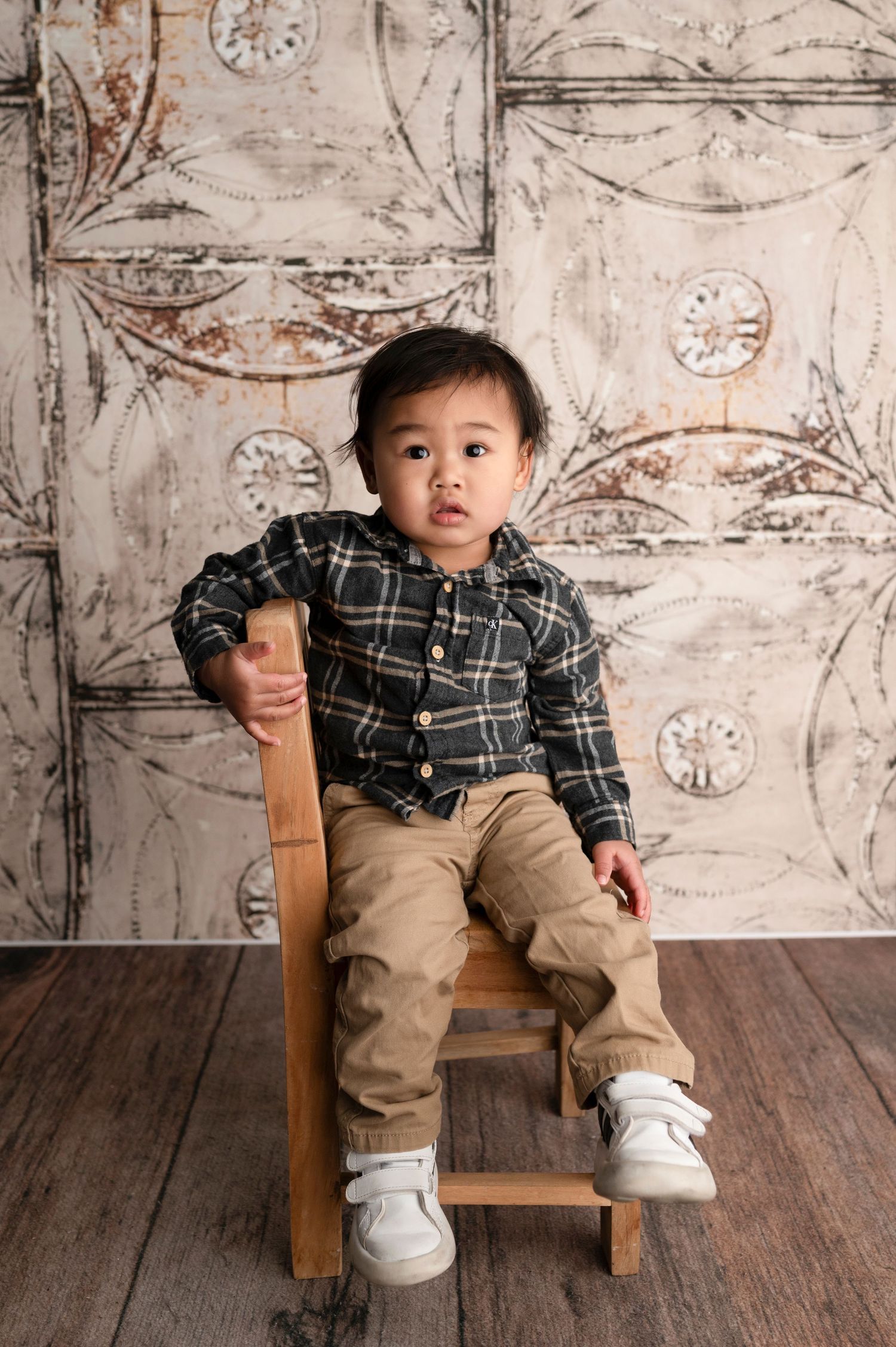Toddler boy in plaid shirt and khaki pants sitting on small wooden chair against decorative white backdrop.