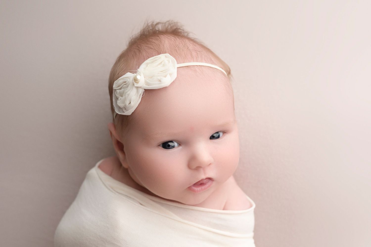 Newborn baby wrapped in white swaddle cloth wearing a delicate white flower headband, posed against a neutral background.
