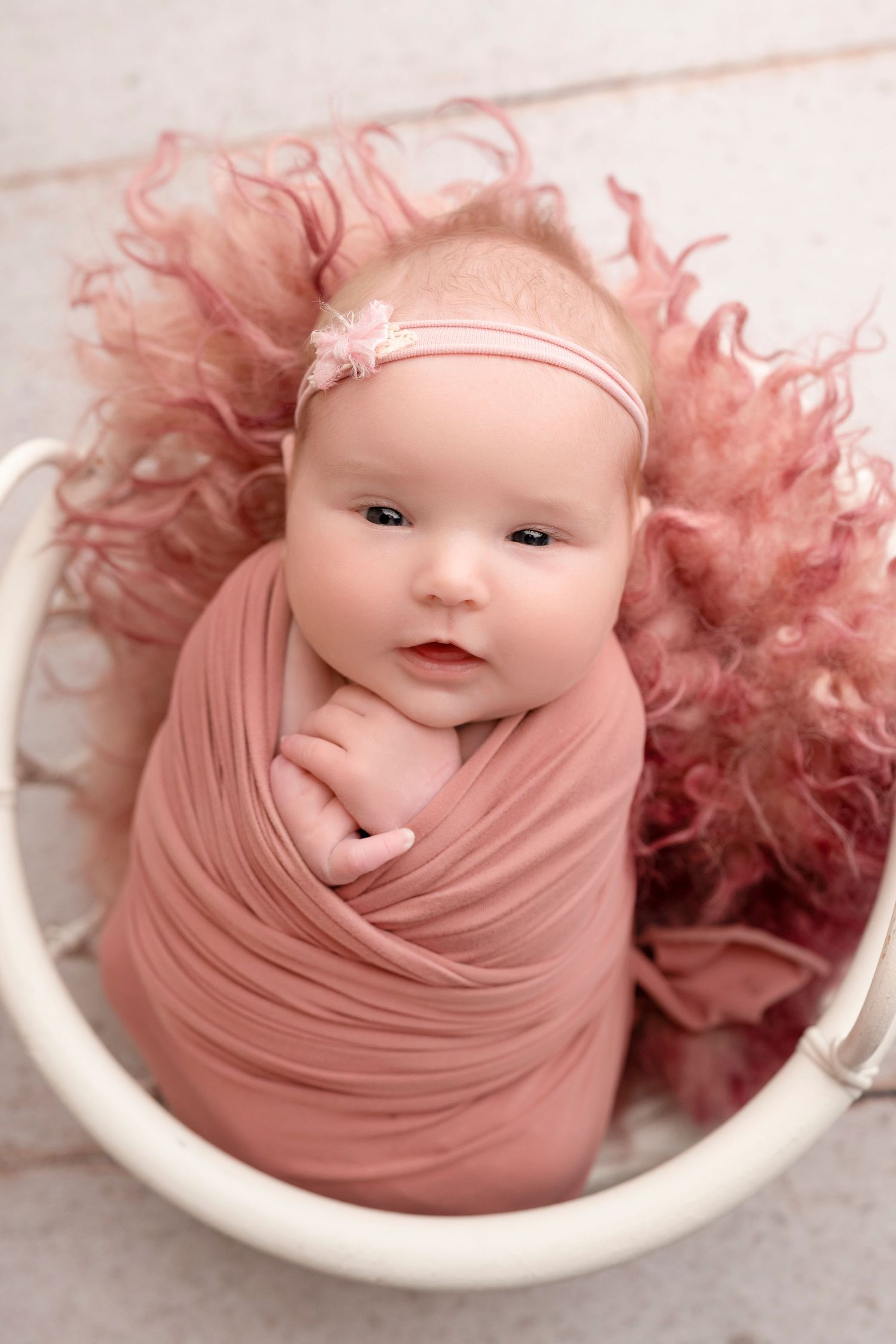 Newborn baby girl swaddled in pink wrap, wearing floral headband, nestled in white bucket with fluffy pink fur.