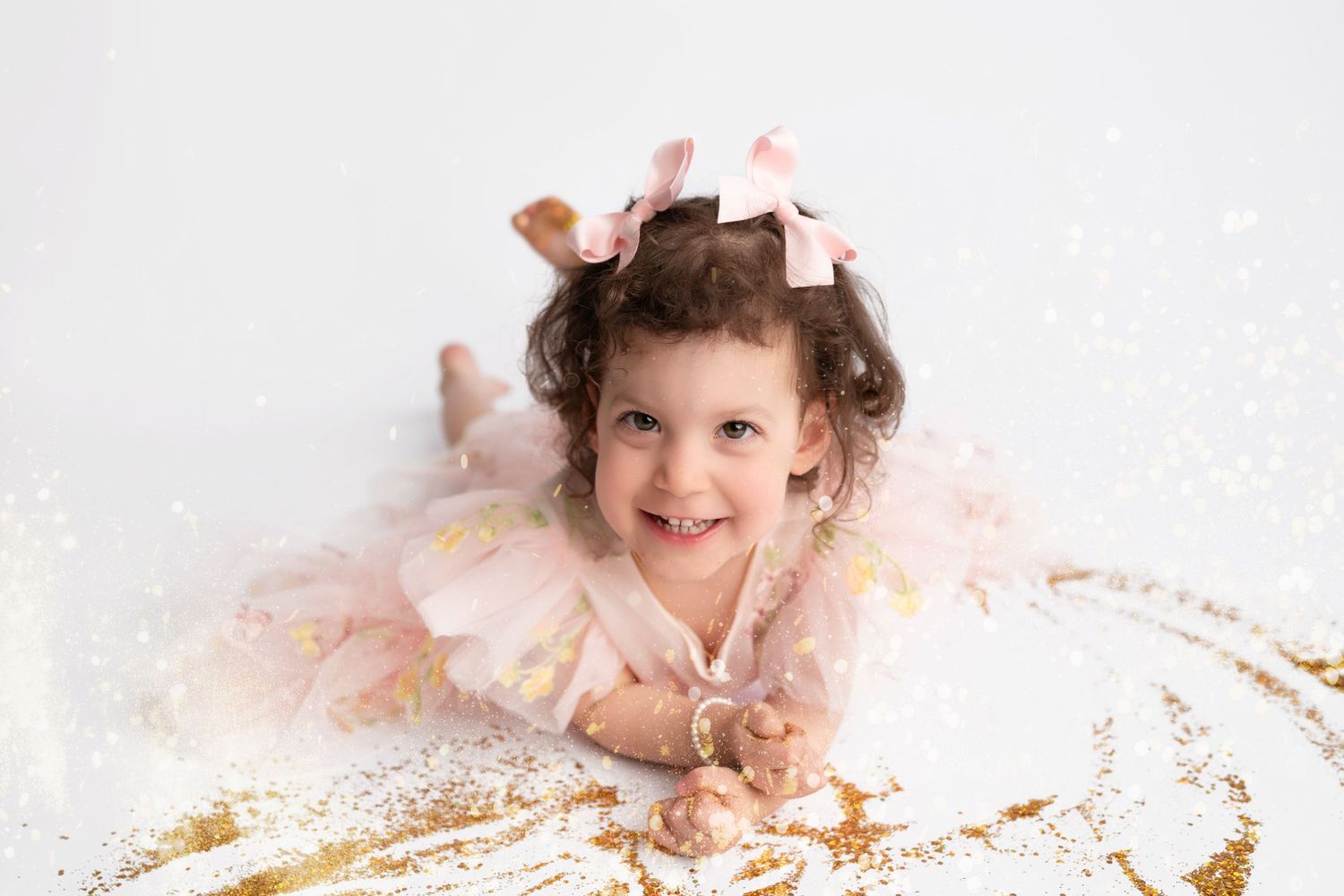 Child in white dress lying on floor surrounded by pink and gold glitter creates joyful moment during photo session.