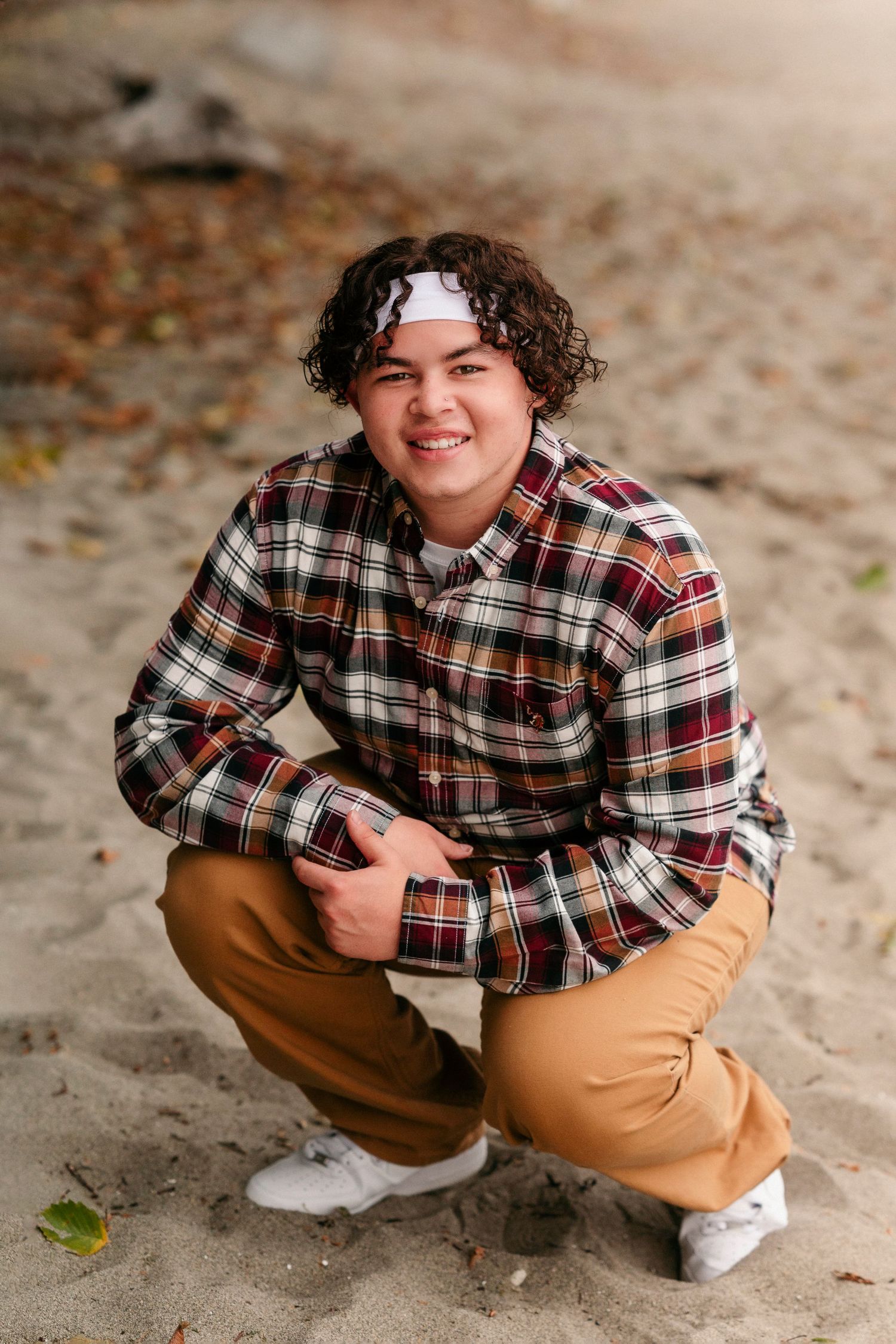 Person in plaid shirt and khaki pants crouches on sandy ground in outdoor portrait session.