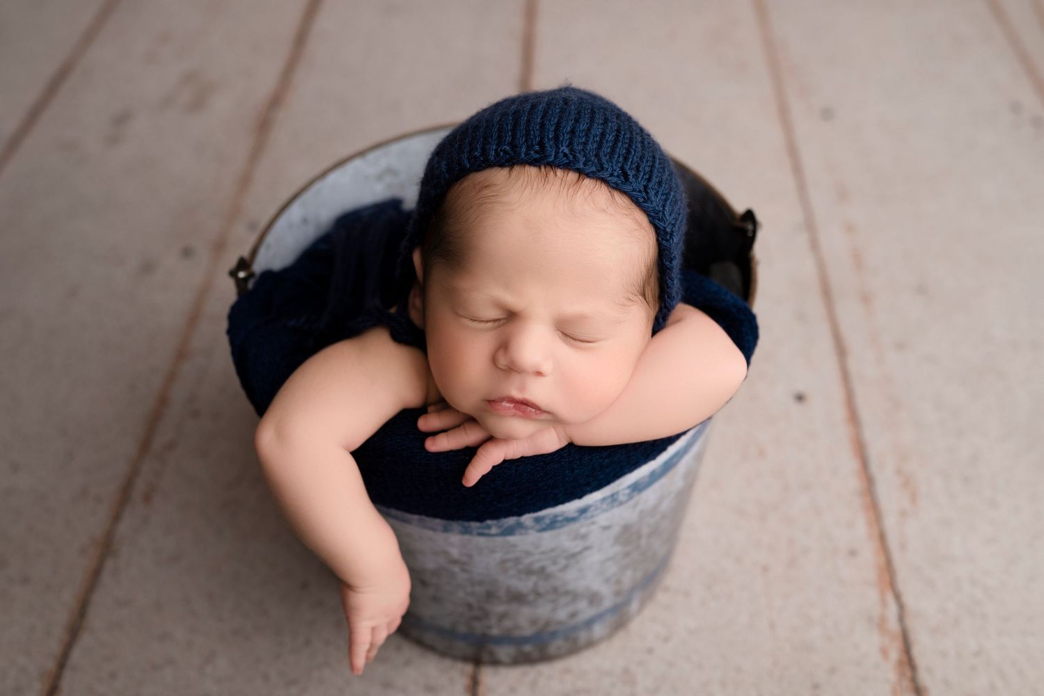 Sleeping newborn nestled in dark blue knit hat inside a weathered metal bucket.