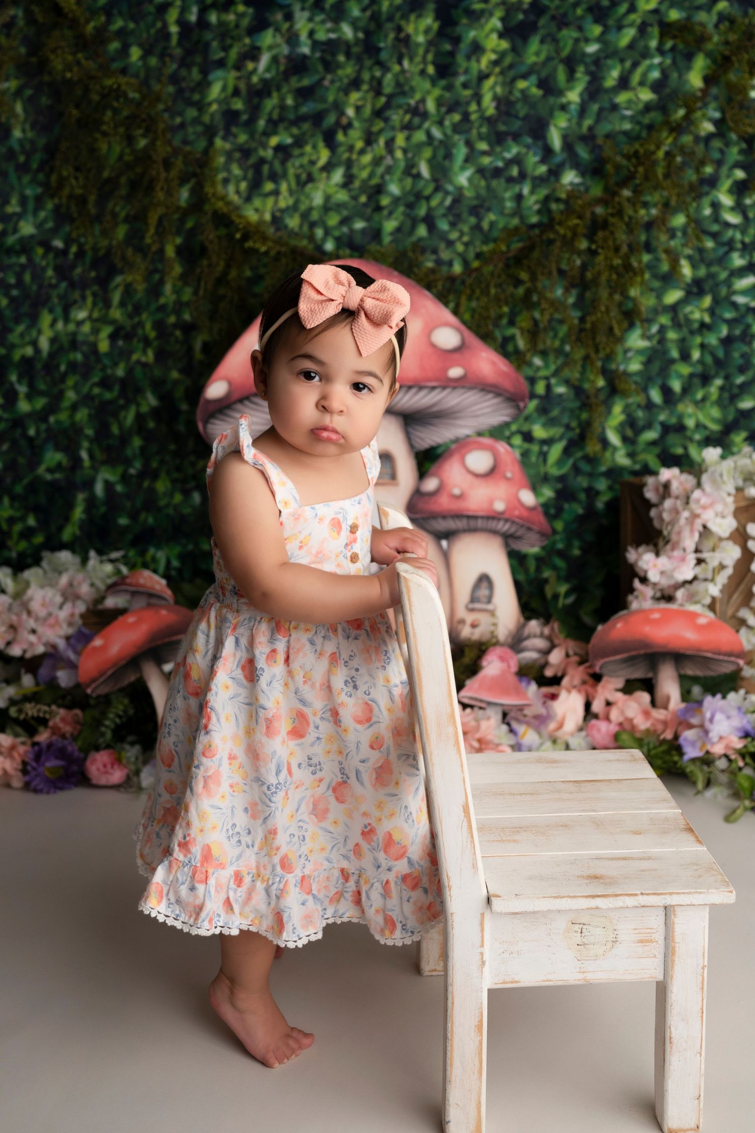 Baby in floral dress poses with whimsical red mushroom decorations and white wooden bench in garden-themed setting.