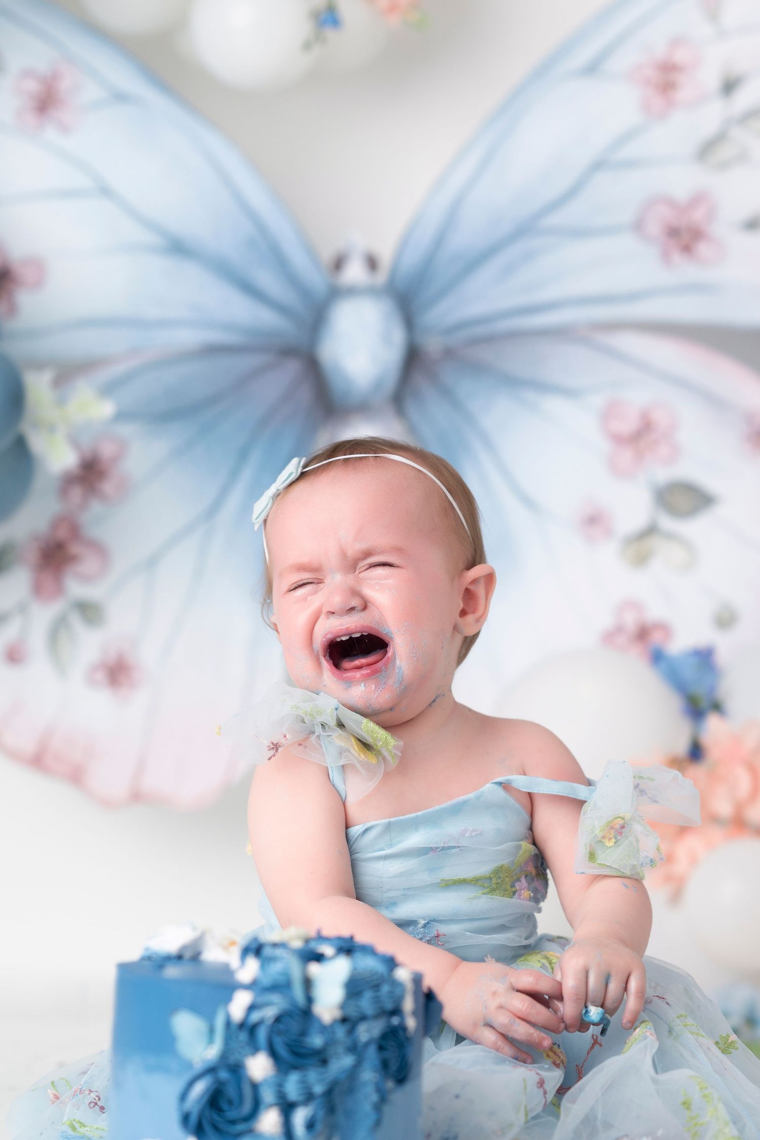 Baby wearing blue dress laughs joyfully on floral background with butterfly decorations in studio setting.