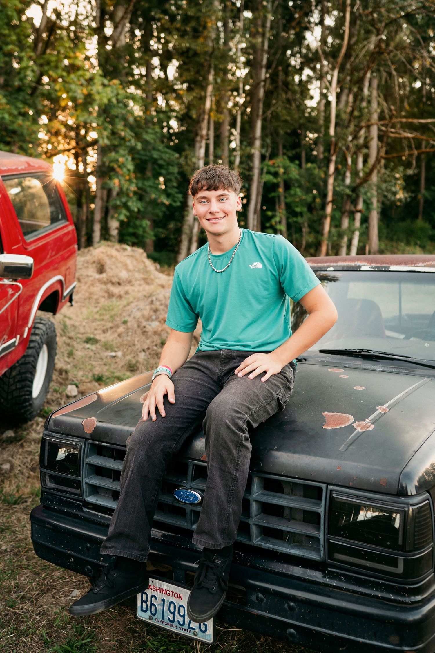 Someone poses sitting on the hood of a black vintage car in a wooded setting during golden hour.