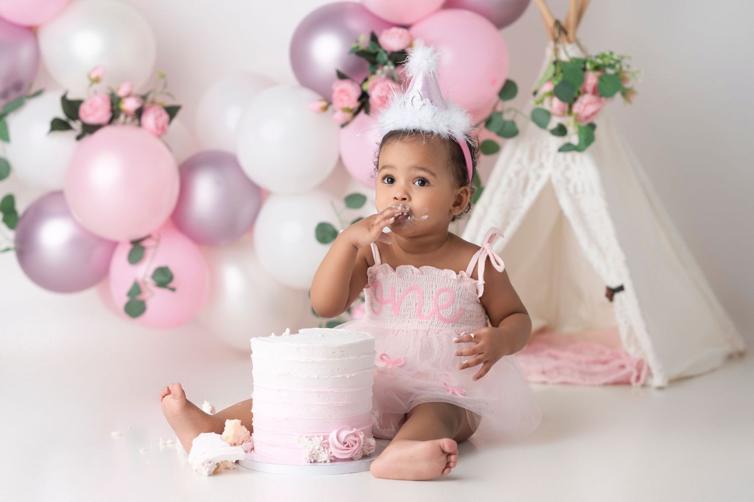 Baby girl in pink romper eating cake at first birthday smash cake session with balloons and teepee.