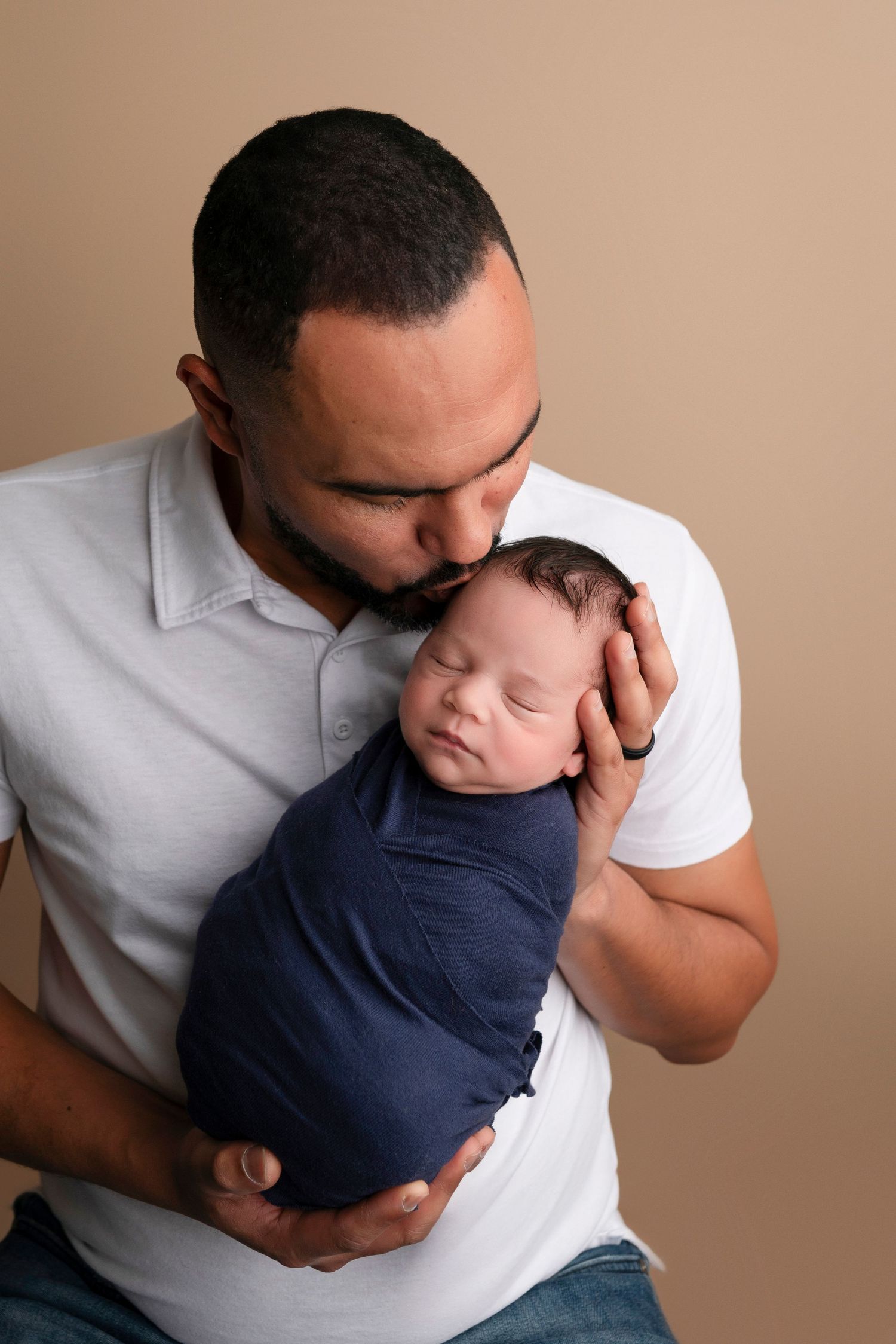 A father tenderly kisses his swaddled newborn baby wrapped in navy blue cloth against a beige background.