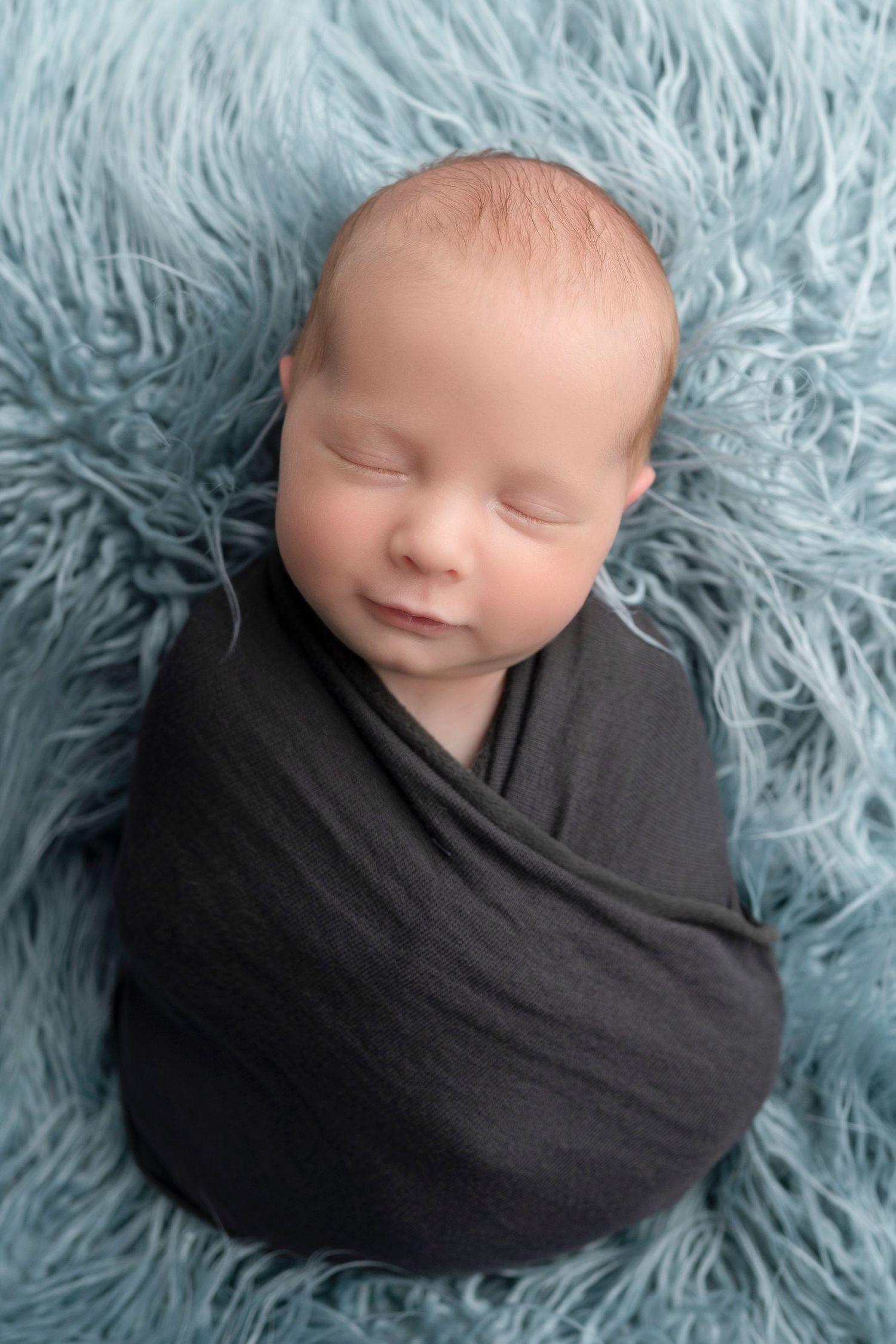 Sleeping newborn baby swaddled in dark gray wrap on a soft blue fluffy rug, peaceful smile.