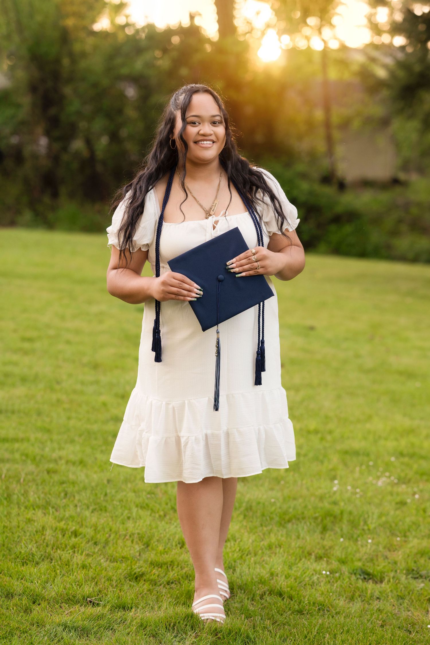 Graduate in white dress holds diploma while walking on grass at sunset during outdoor graduation photoshoot.