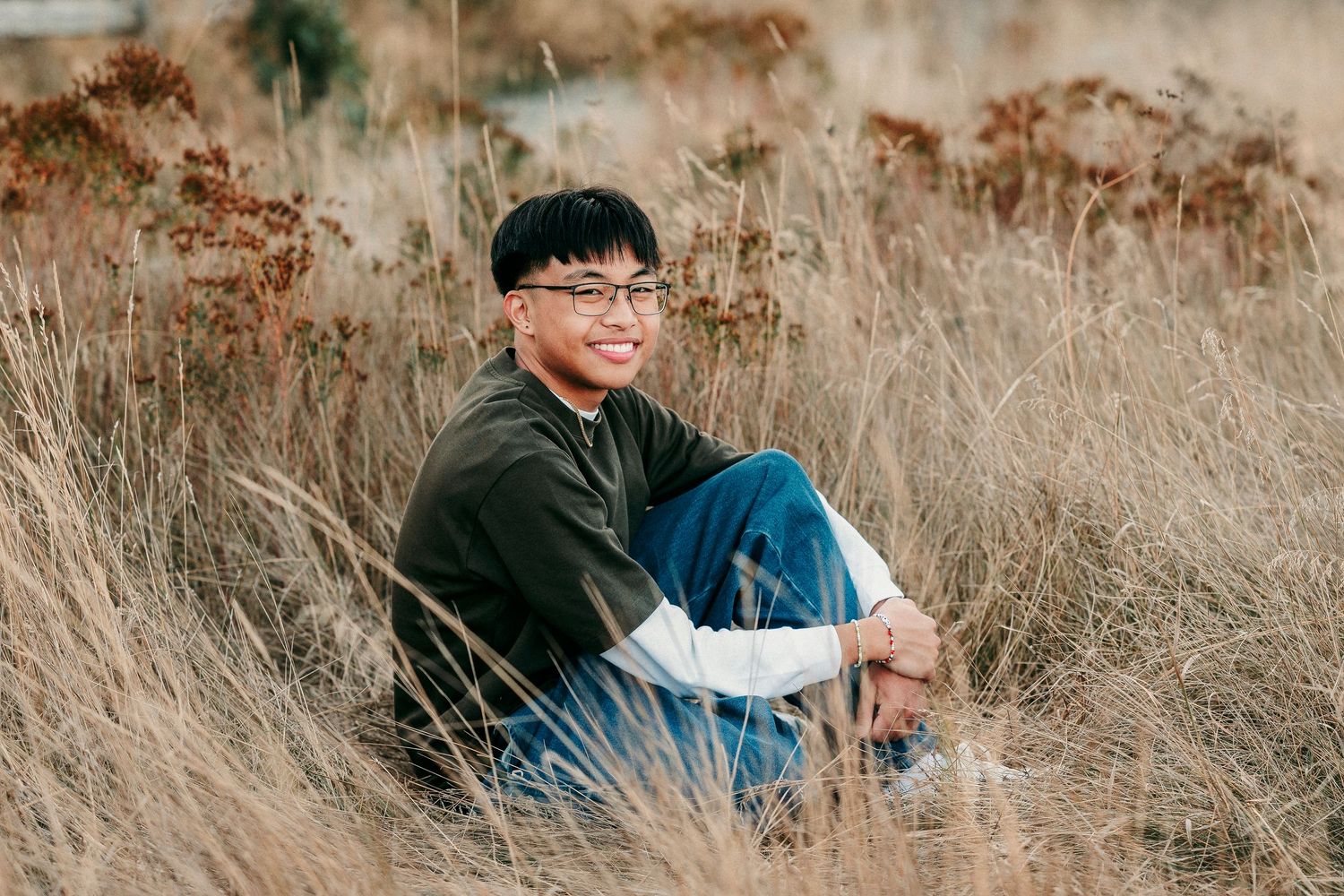 Person sitting in tall brown grass field during autumn season wearing casual clothing and glasses.