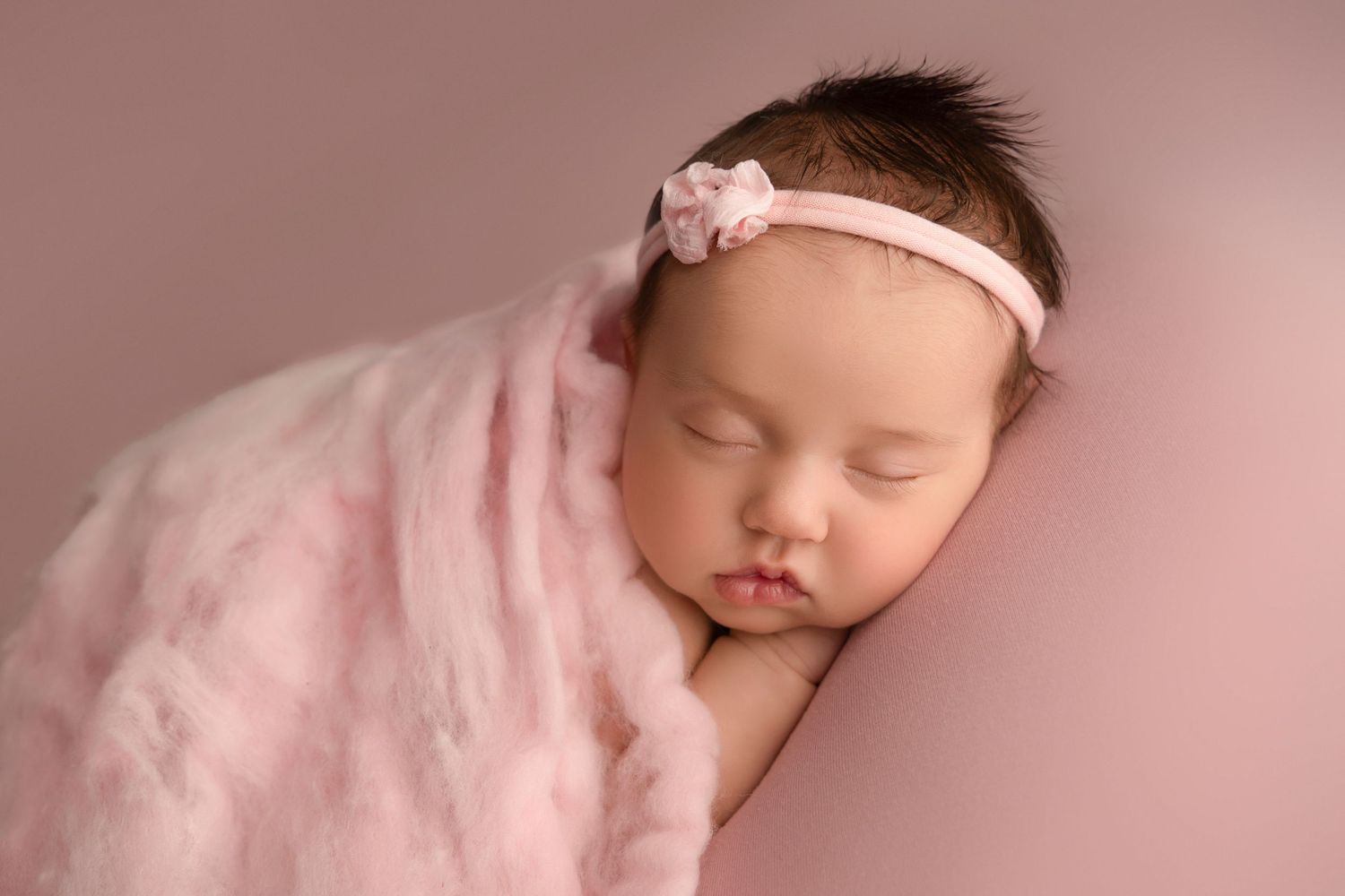 A sleeping newborn baby wearing a pink headband lies peacefully on soft pink blankets in a professional photo setting.
