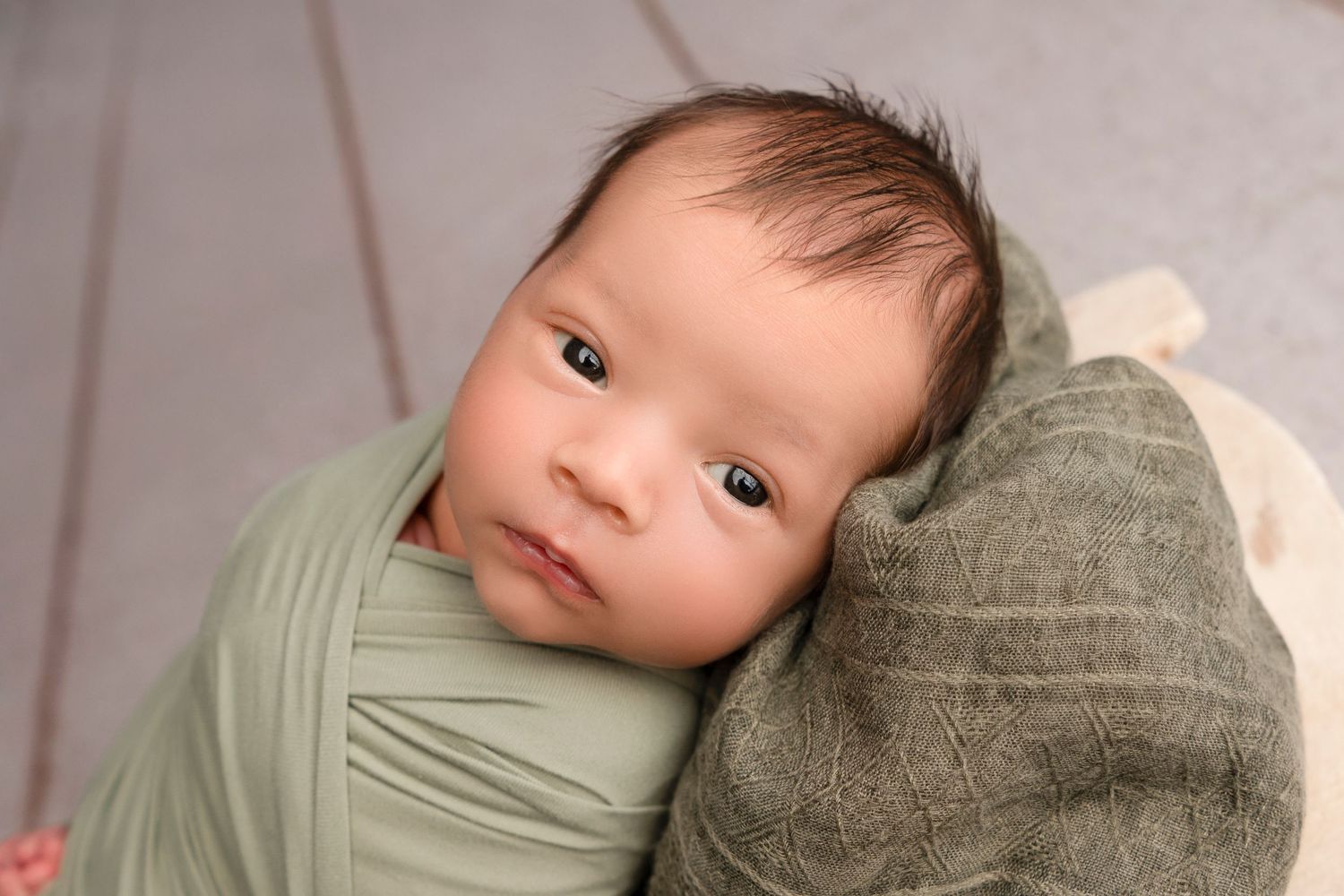 A sleeping baby wrapped in soft sage green fabric rests peacefully on a textured gray cushion.