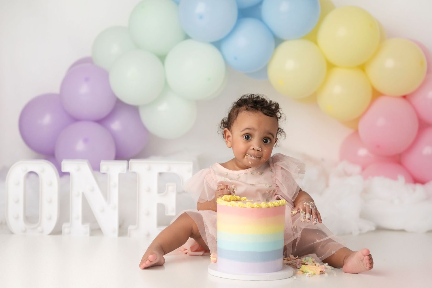 Baby in pastel rainbow cake smash photo session with balloon arch backdrop and white ONE letters.
