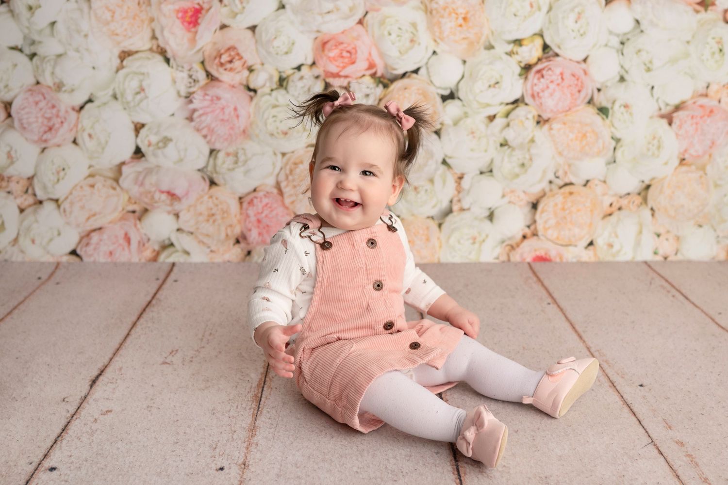Smiling baby girl in pink overalls sits on wooden floor against a floral backdrop of white and pink roses.