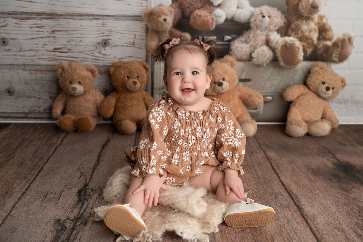 Smiling baby girl in floral brown outfit sits on fur rug surrounded by teddy bears on rustic wood floor.