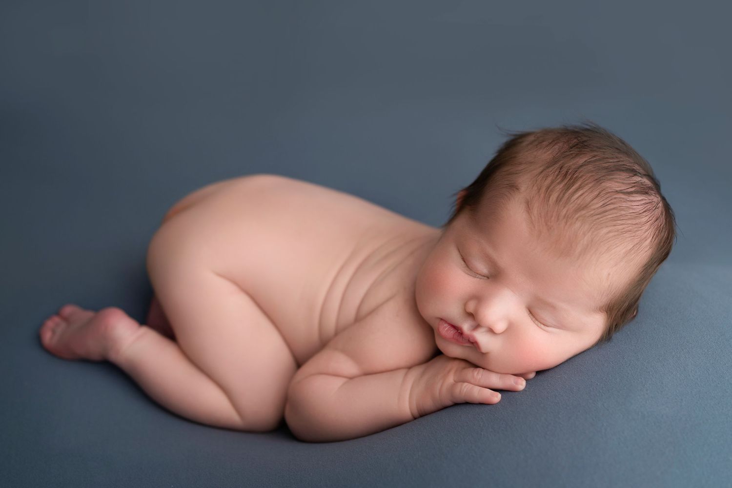Peaceful newborn baby sleeping curled up on a soft blue-gray backdrop in a classic newborn photography pose.
