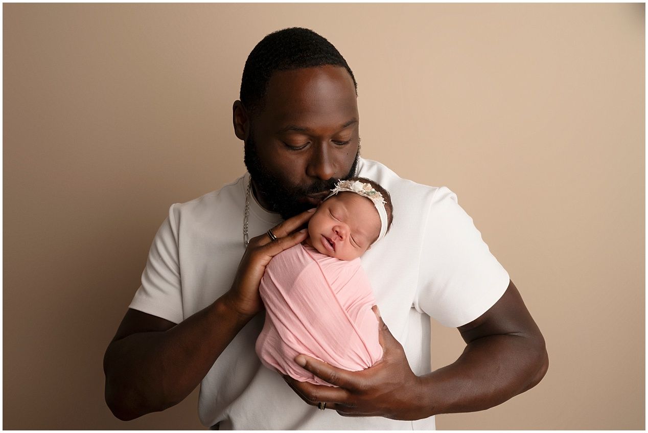 A father in a white t-shirt tenderly kisses his newborn baby wrapped in a pink swaddle against a beige background.