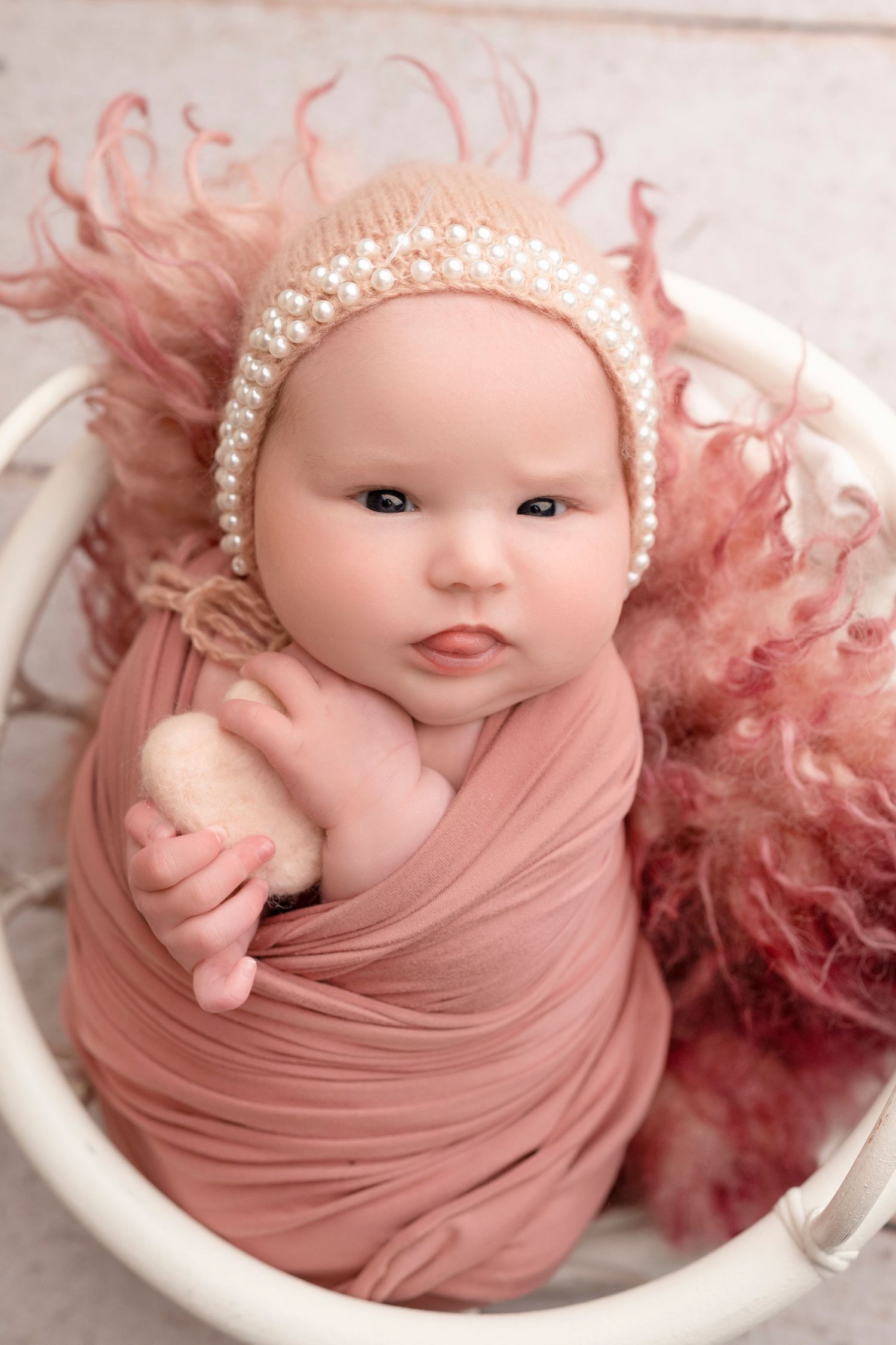 Newborn baby wrapped in pink swaddle with pearl bonnet, nestled in white basket with fluffy pink fur.