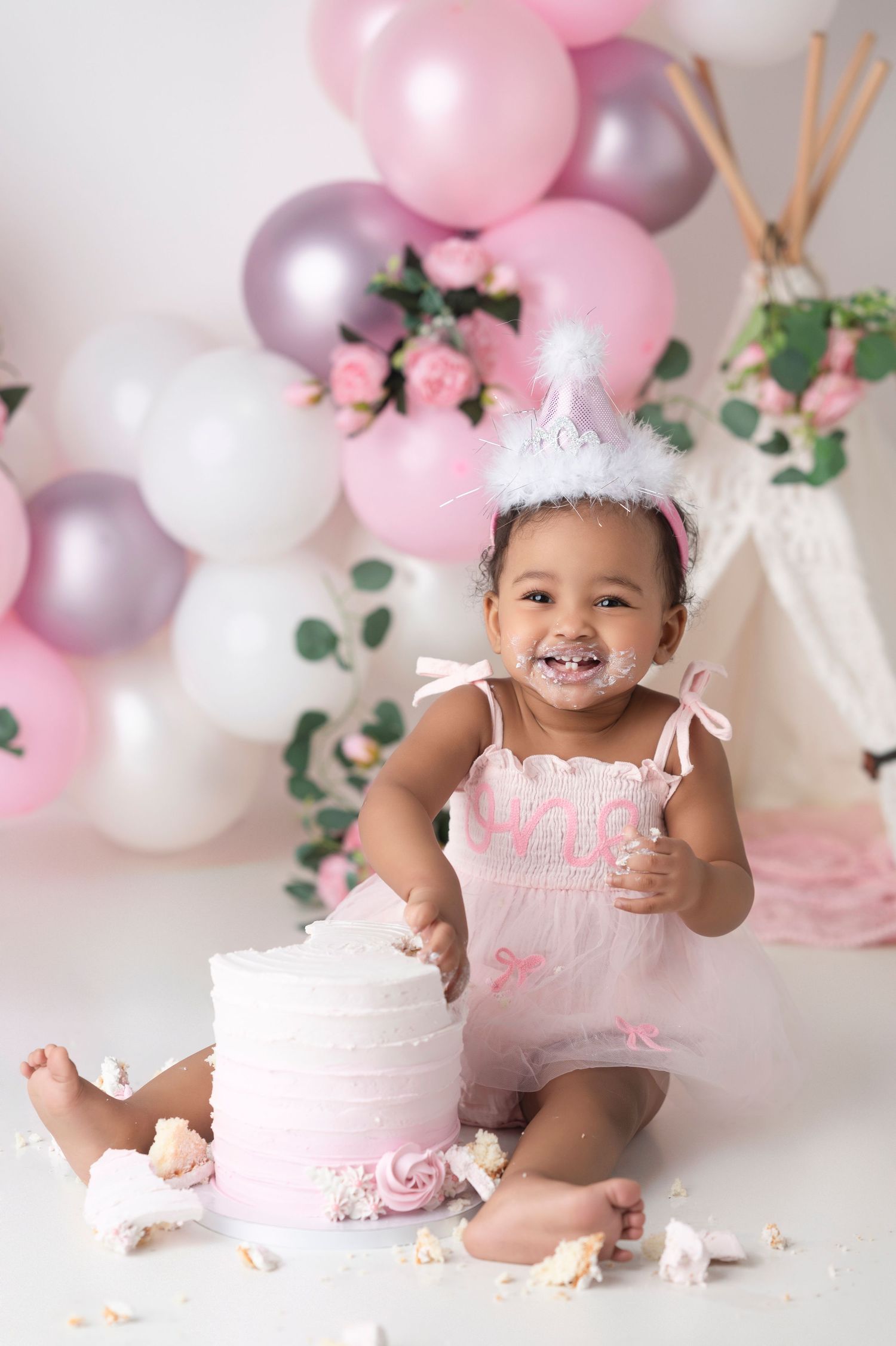 Smiling baby girl in pink dress and crown celebrates first birthday smashing a pink cake with balloons behind her.