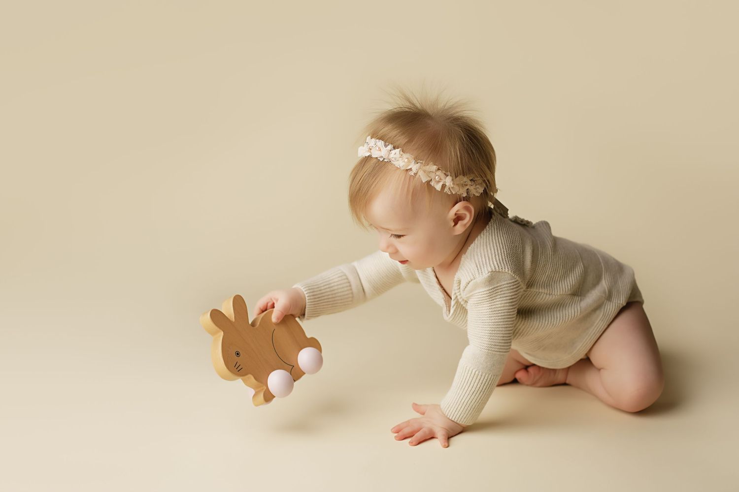 Baby wearing white headband and onesie crawls toward a gingerbread cookie on cream-colored backdrop.