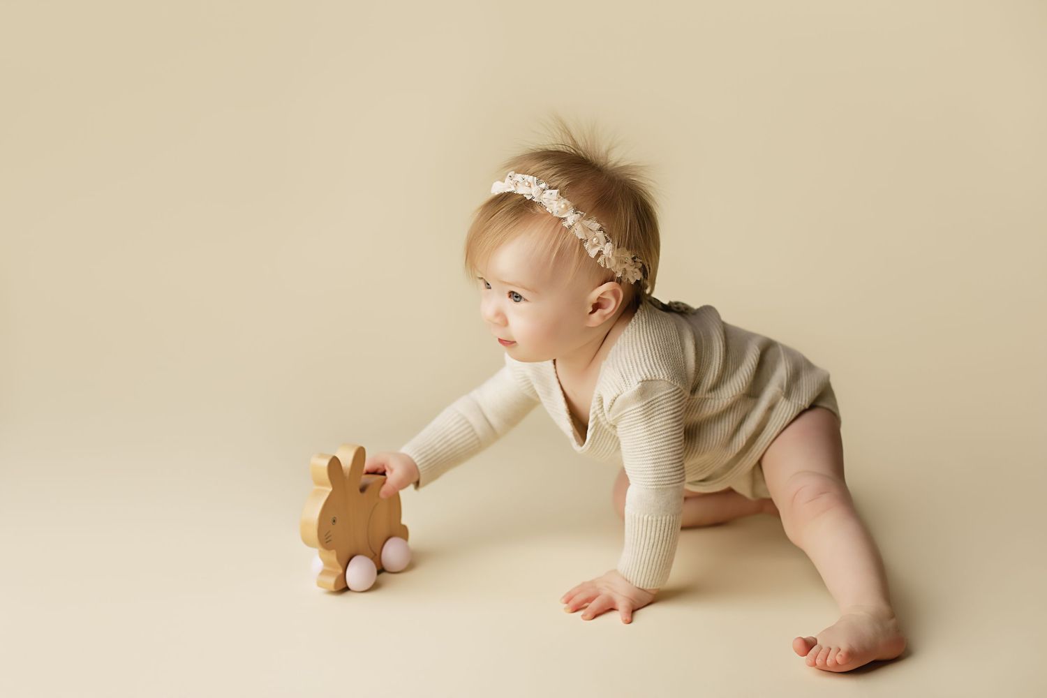 A baby in a white onesie reaches out to touch a small plush toy bear on a cream-colored background.