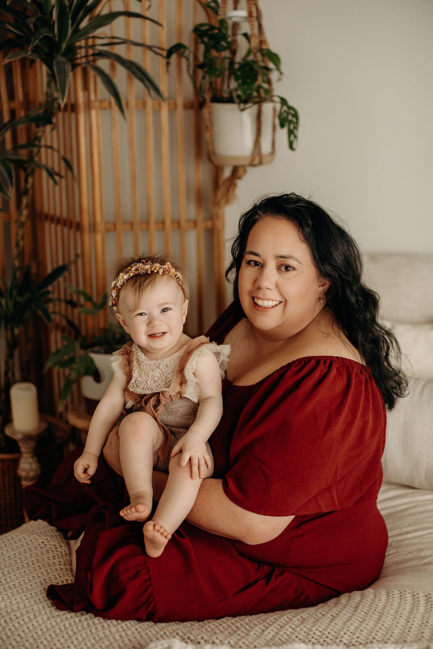Mother in red dress and child in lace outfit sitting on floor with indoor plants and bamboo decor.