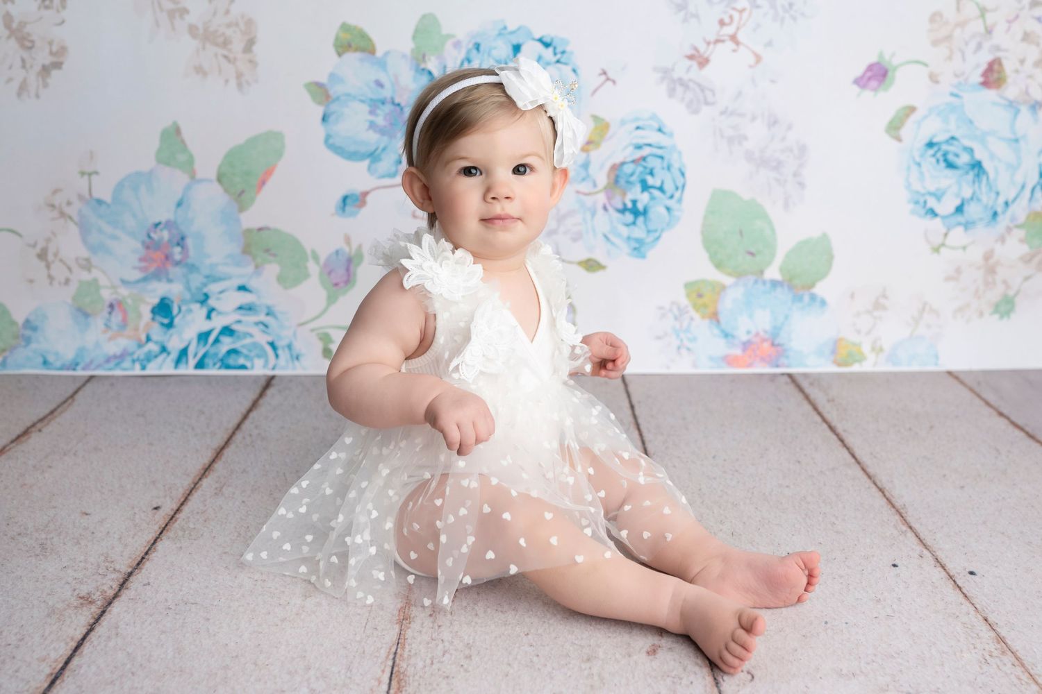 Baby girl in white polka dot tulle dress sits on wood floor against floral backdrop for first birthday photo.