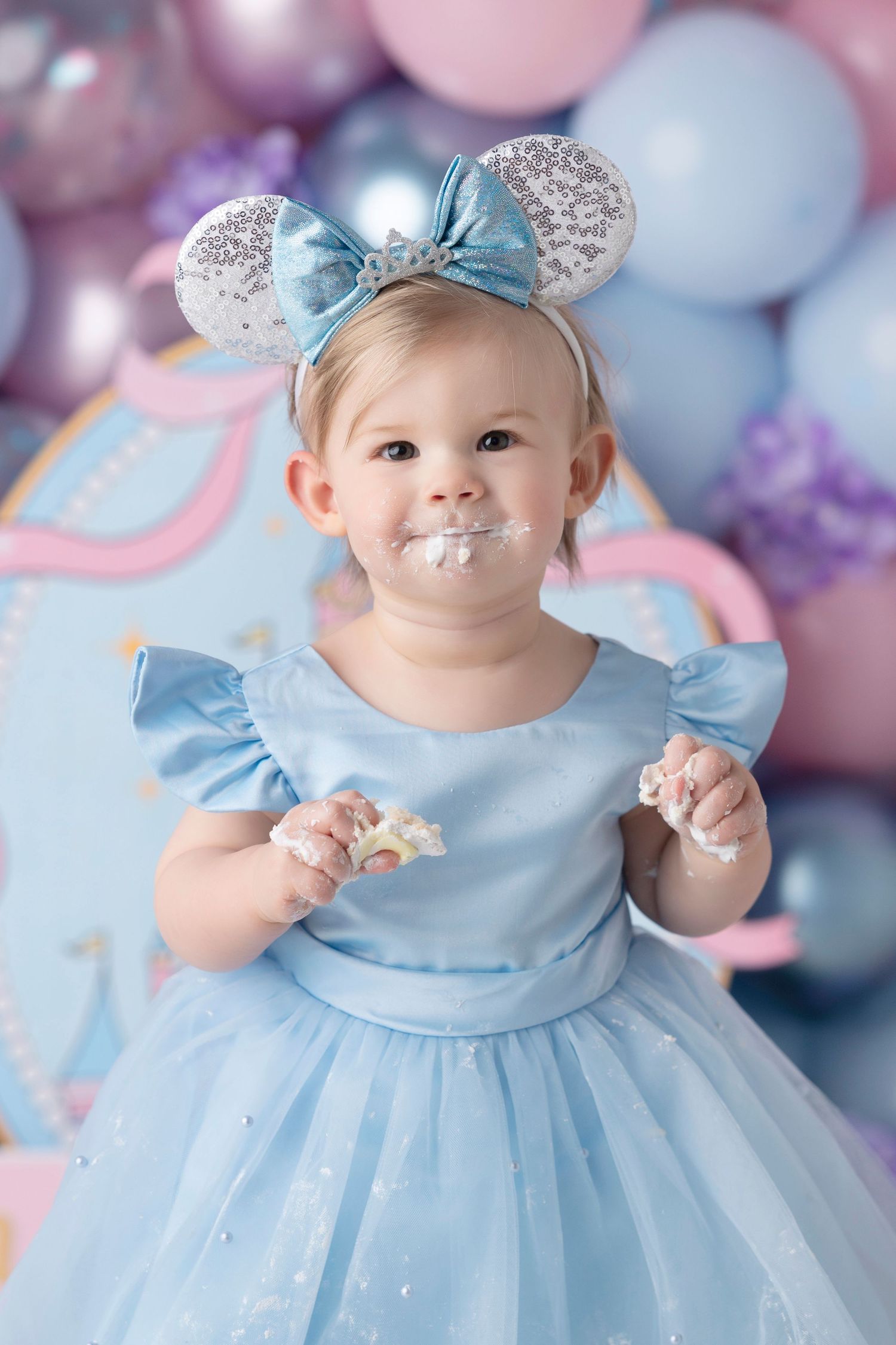 Toddler girl in blue Cinderella dress with Minnie Mouse ears eating birthday cake smash with balloons backdrop.