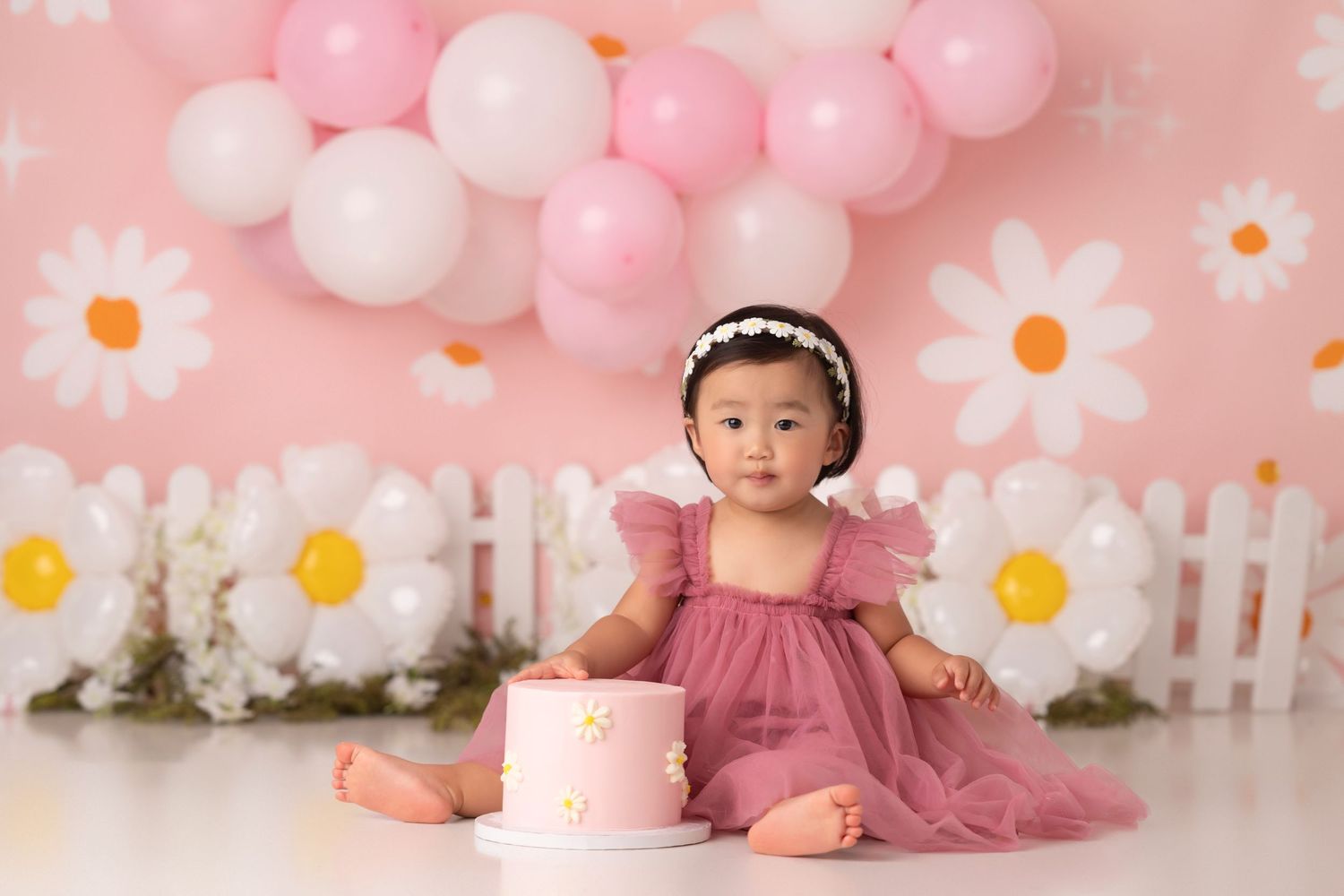 Baby girl in pink dress sits beside a daisy cake, surrounded by pink balloons and daisy decorations for her first birthday.