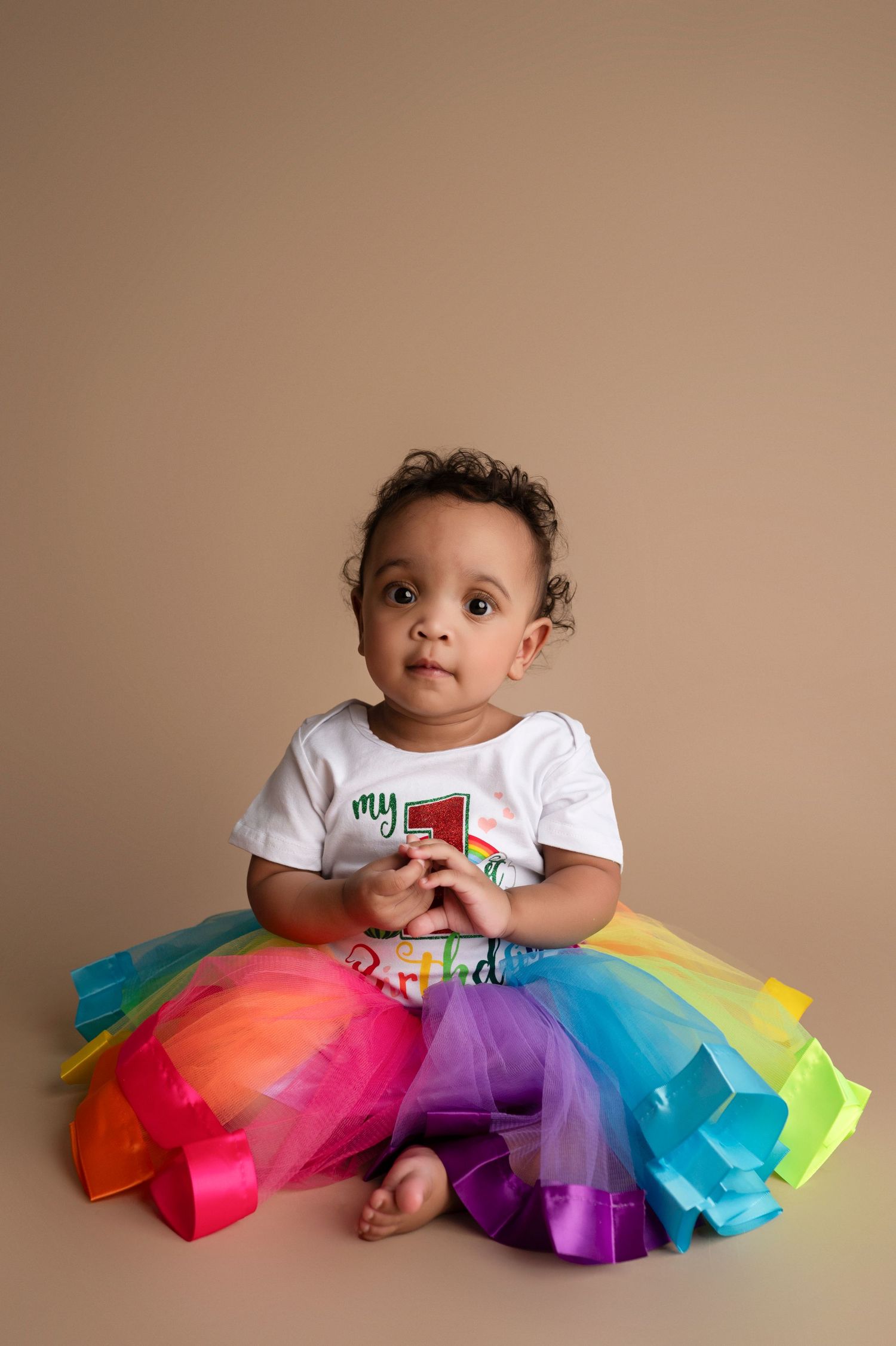 Toddler wearing white t-shirt and colorful rainbow tutu sits against neutral beige background during portrait session.