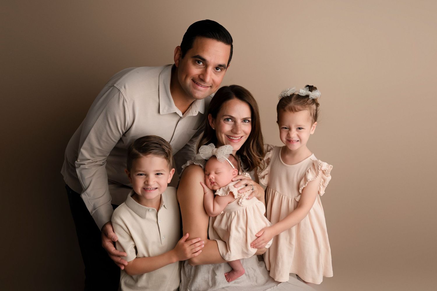 A family of five poses together in coordinated neutral-colored outfits against a beige studio backdrop.