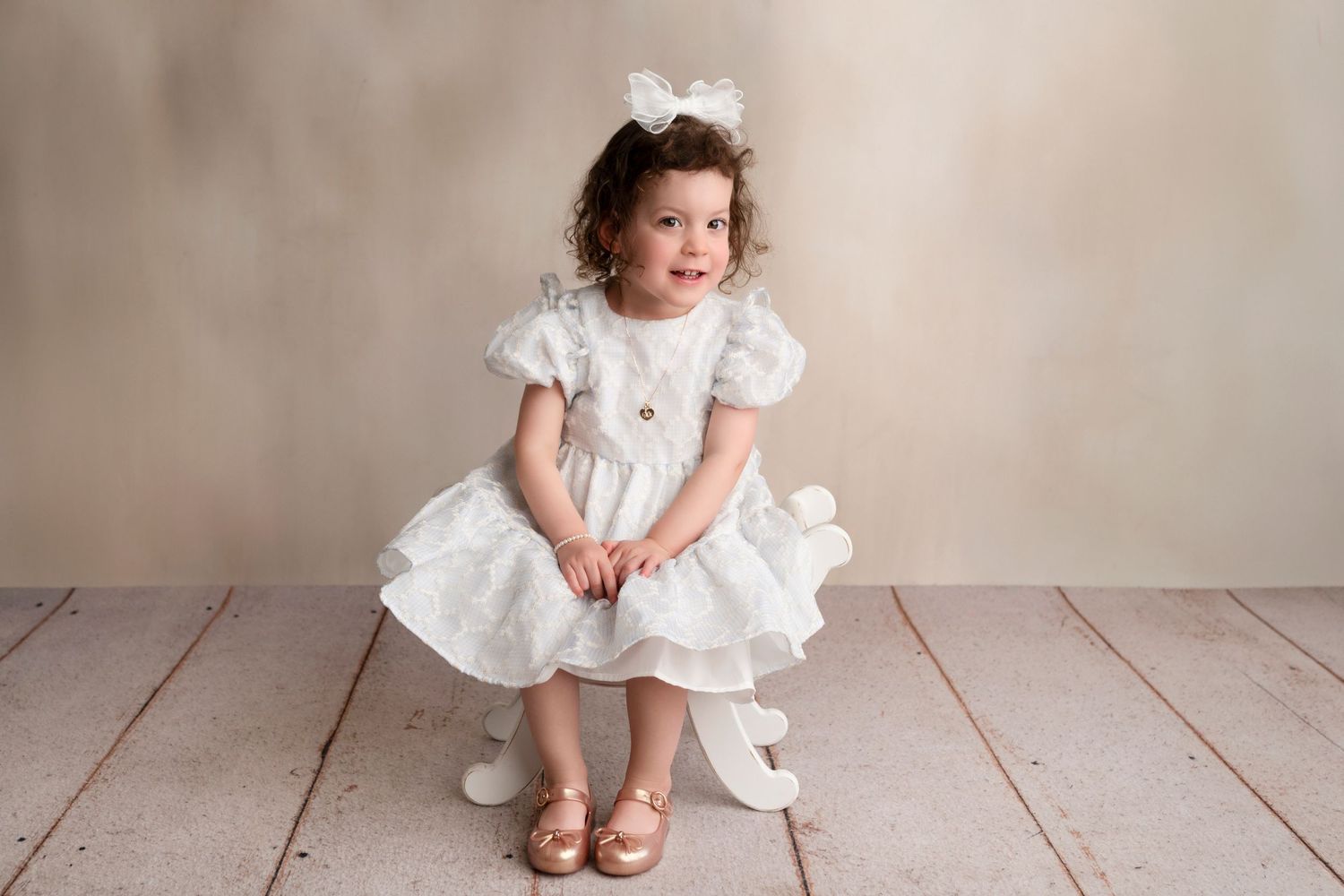 A small child in a white ruffled dress and crown sits on a wooden floor against a neutral backdrop.