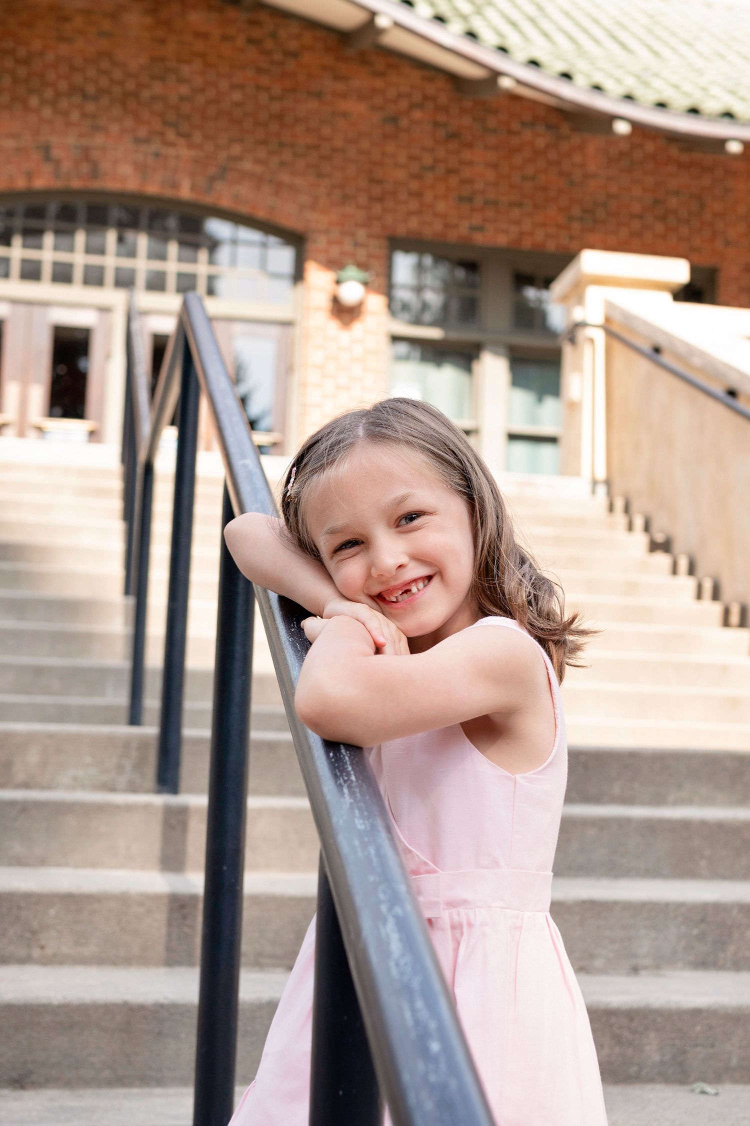 A young child in a pink dress smiles playfully while leaning against a metal railing near brick steps.