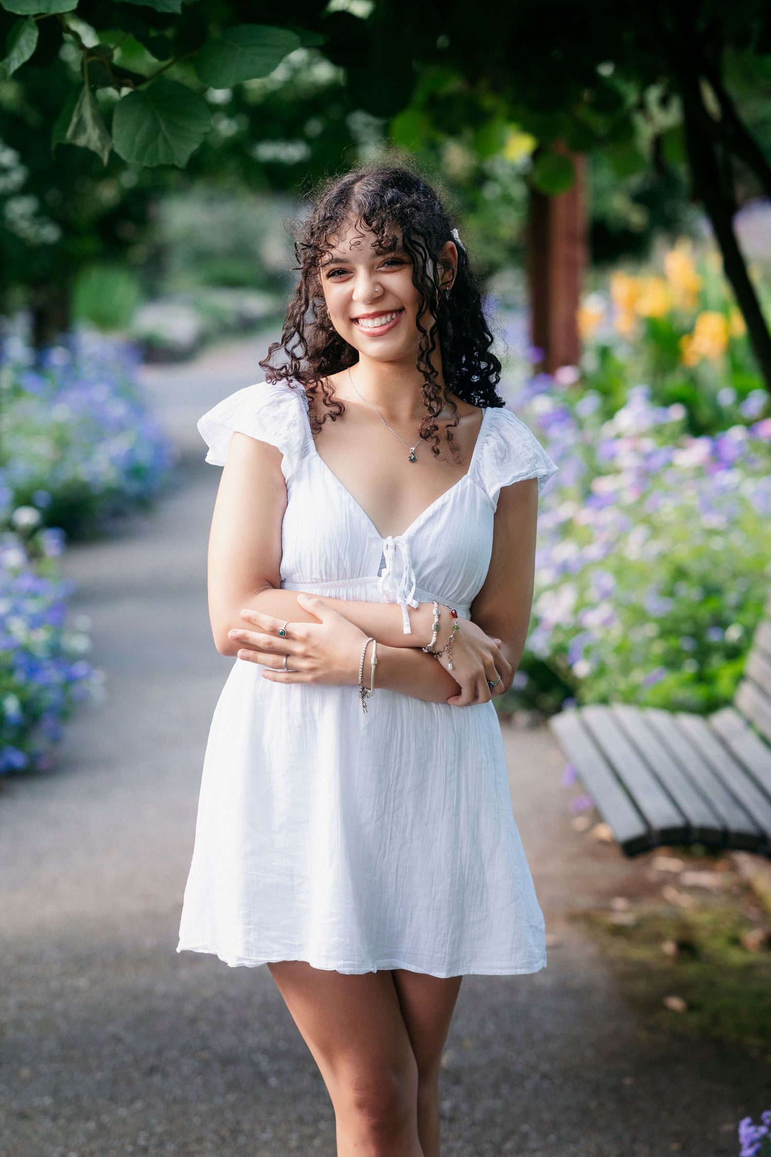 A person in a flowing white summer dress poses playfully in a garden setting with colorful flowers in the background.