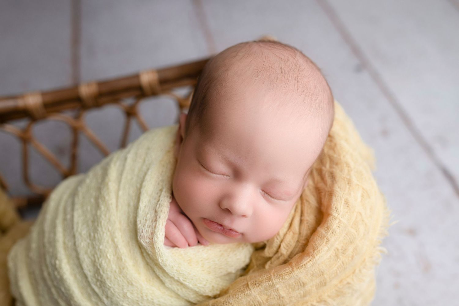 Newborn baby swaddled in a soft yellow wrap, sleeping peacefully in a wicker basket on a white wooden floor.