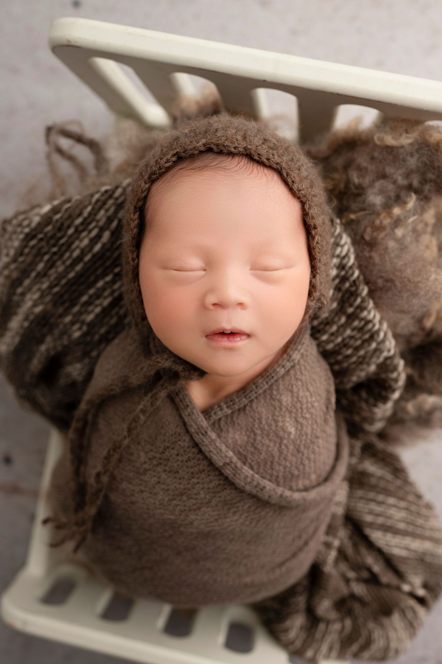 Newborn baby swaddled in brown knit wrap wearing a bonnet, sleeping peacefully in a small white crib.