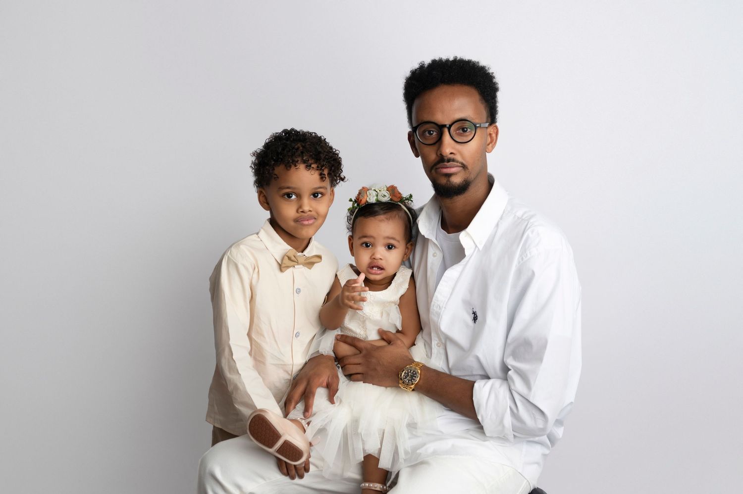 A man in white shirt holds a baby girl while a young boy in bow tie poses beside them for a family portrait.