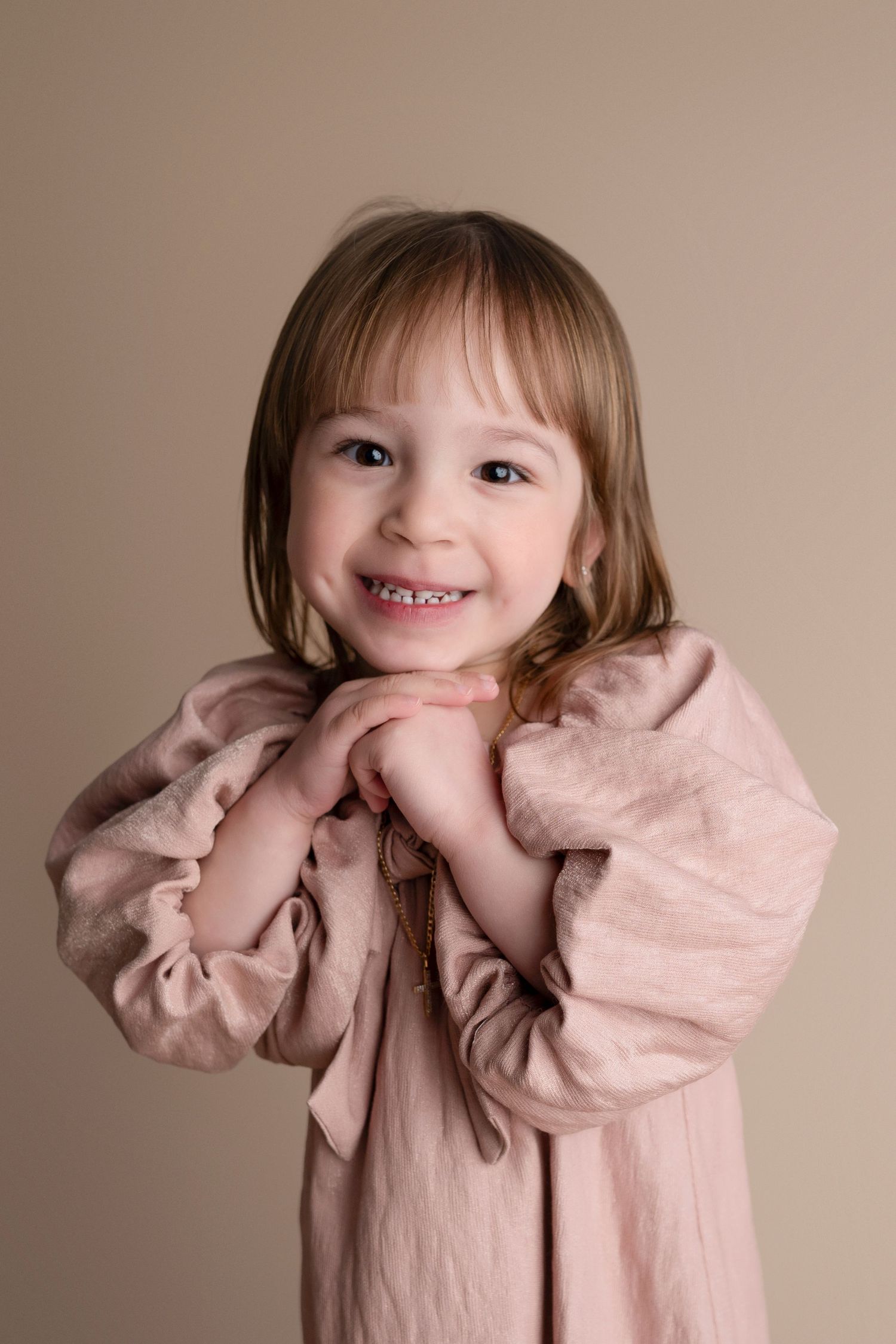 Smiling young girl in a pink dress with hands clasped under chin, posing for a portrait photo.