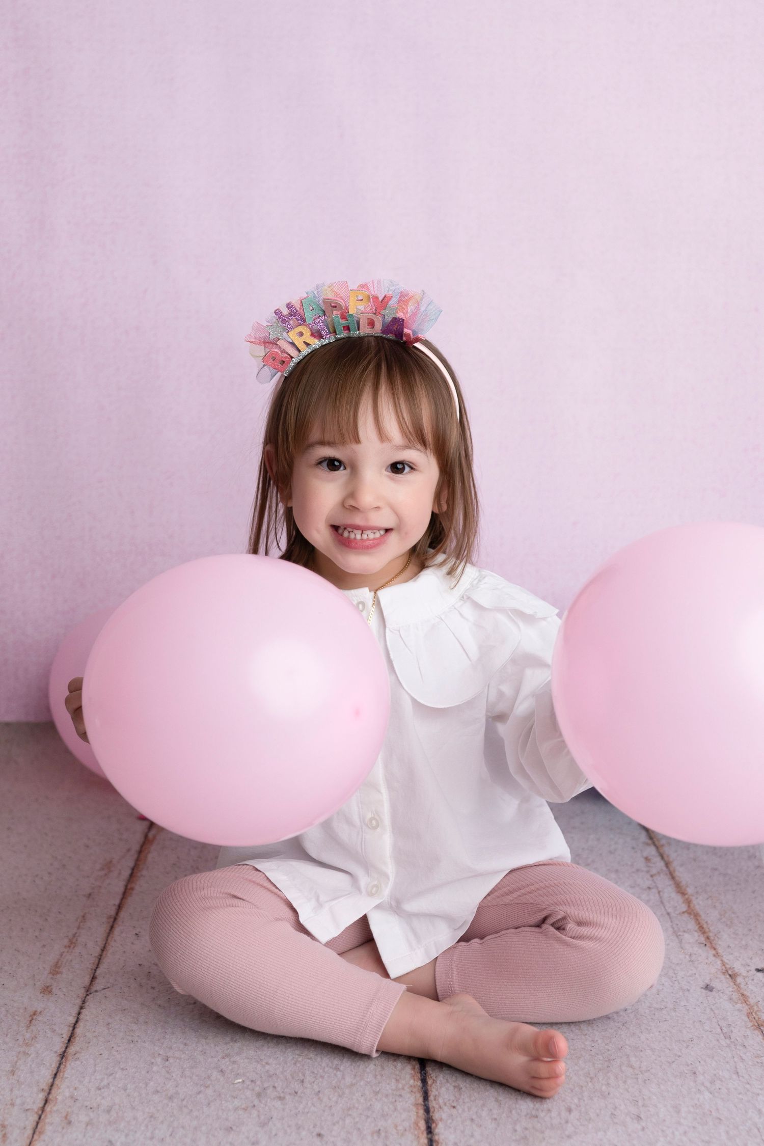 Smiling toddler girl wearing a birthday crown headband, sitting cross-legged with two pink balloons on a pink background.