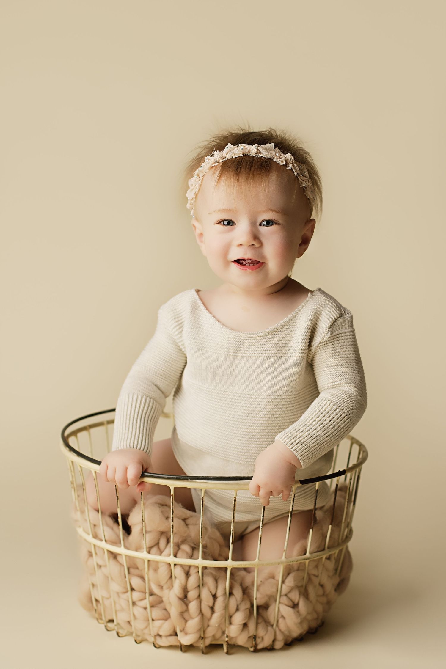 Baby in a knit sweater sits smiling in a woven basket against a neutral beige background.
