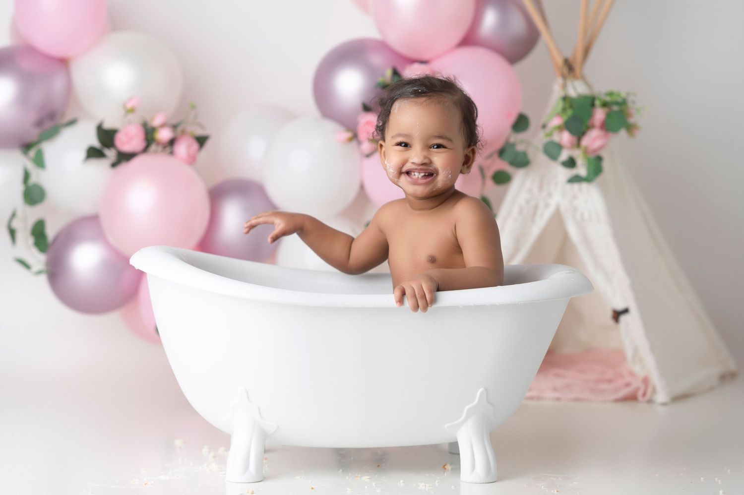 Happy baby sitting in a white clawfoot bathtub surrounded by pink and purple balloons in a studio photo shoot.