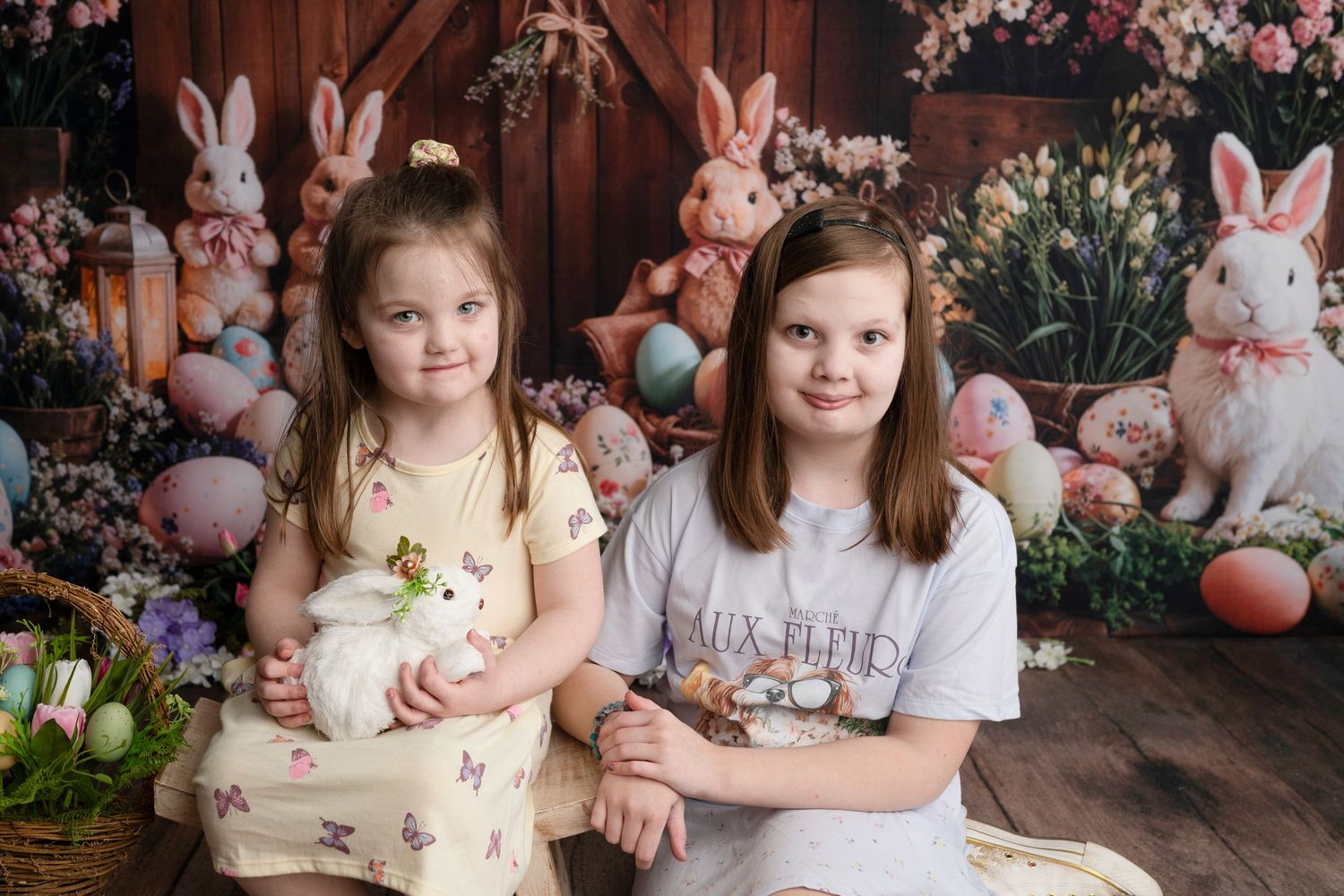 Two young girls pose with a white bunny amid Easter decorations, colorful eggs, and floral backdrop for a holiday portrait.