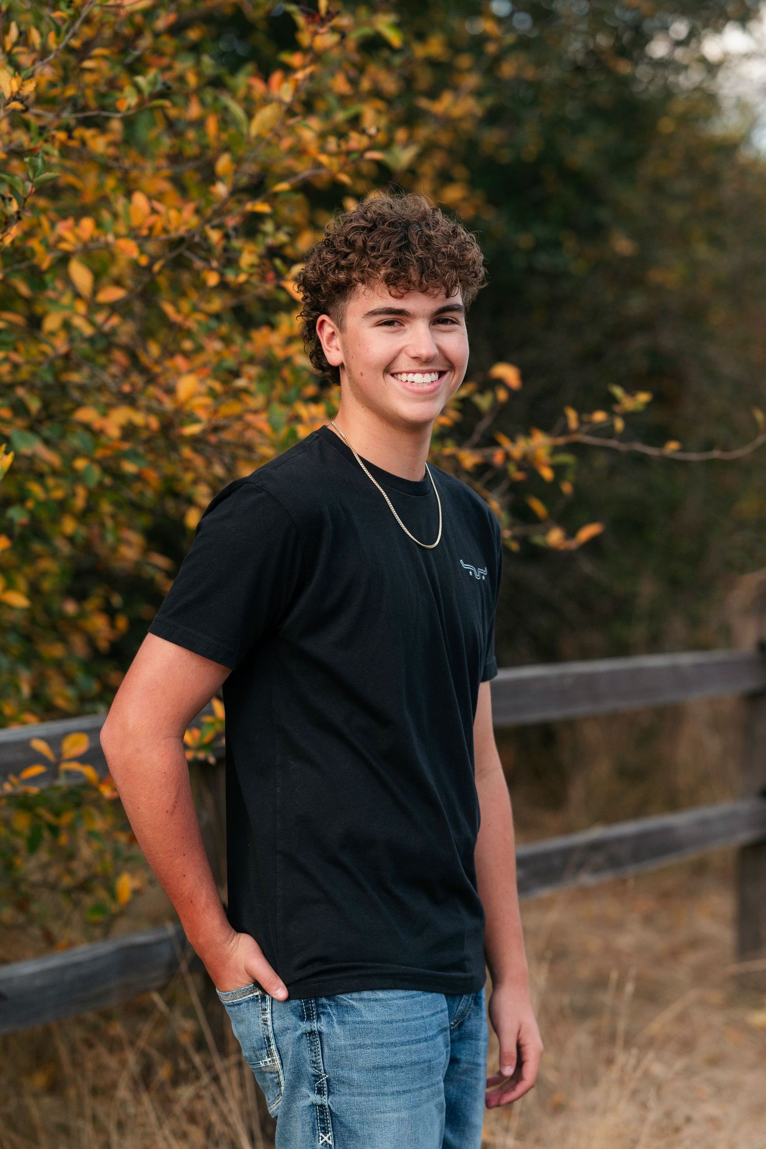 Person in black t-shirt and jeans poses outdoors with autumn foliage in background.