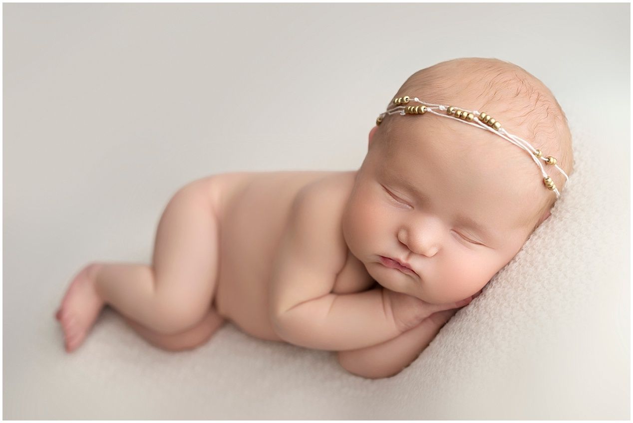 Peaceful sleeping newborn baby wearing delicate headband posed on soft cream backdrop during professional photo session.