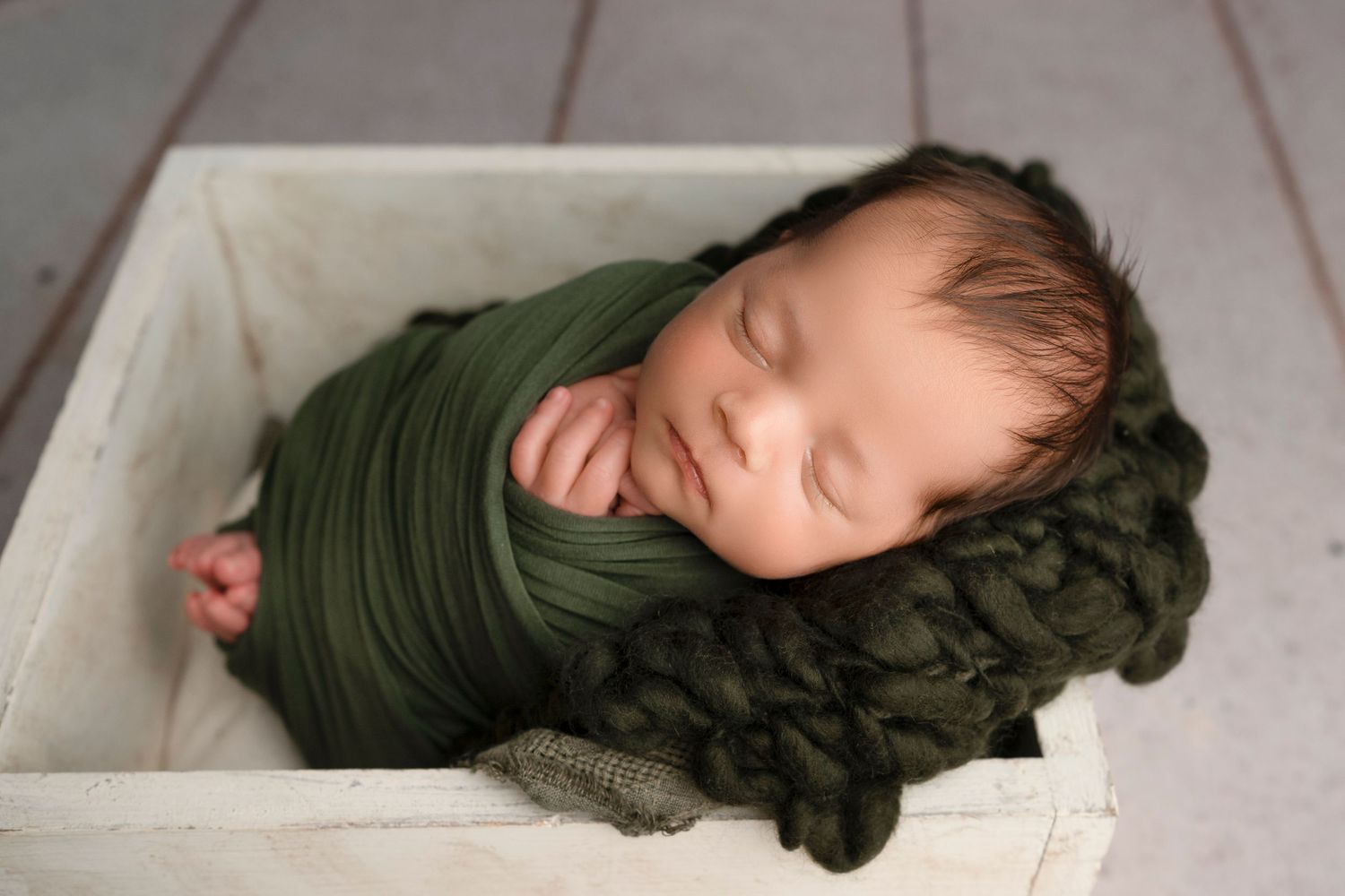 Newborn baby sleeping peacefully wrapped in olive green swaddle blanket on wooden backdrop.